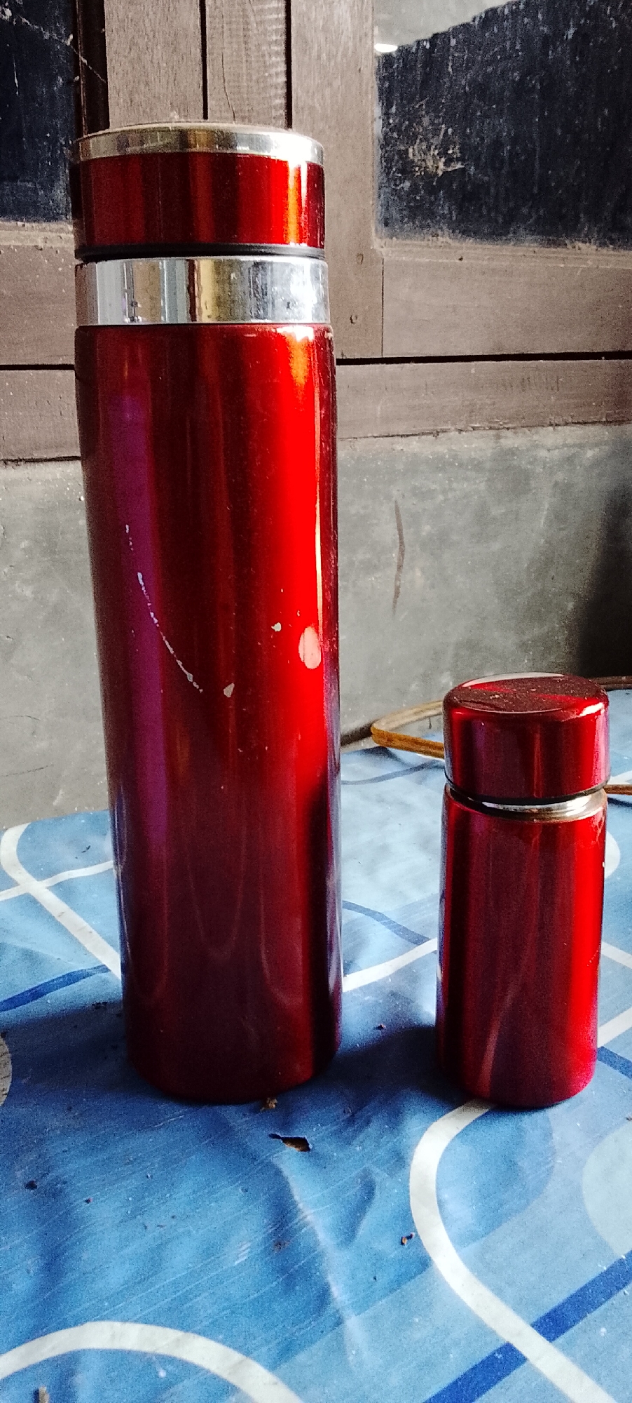 A tall, red, cylindrical thermos stands on a blue and white checkered surface. To its right, a shorter, identical red thermos is also present. Both thermoses have silver bands near their tops. The background features a wooden structure and a grey wall, possibly indicating an indoor or sheltered outdoor setting. The image appears to be taken in Padang Dalom, Indonesia.