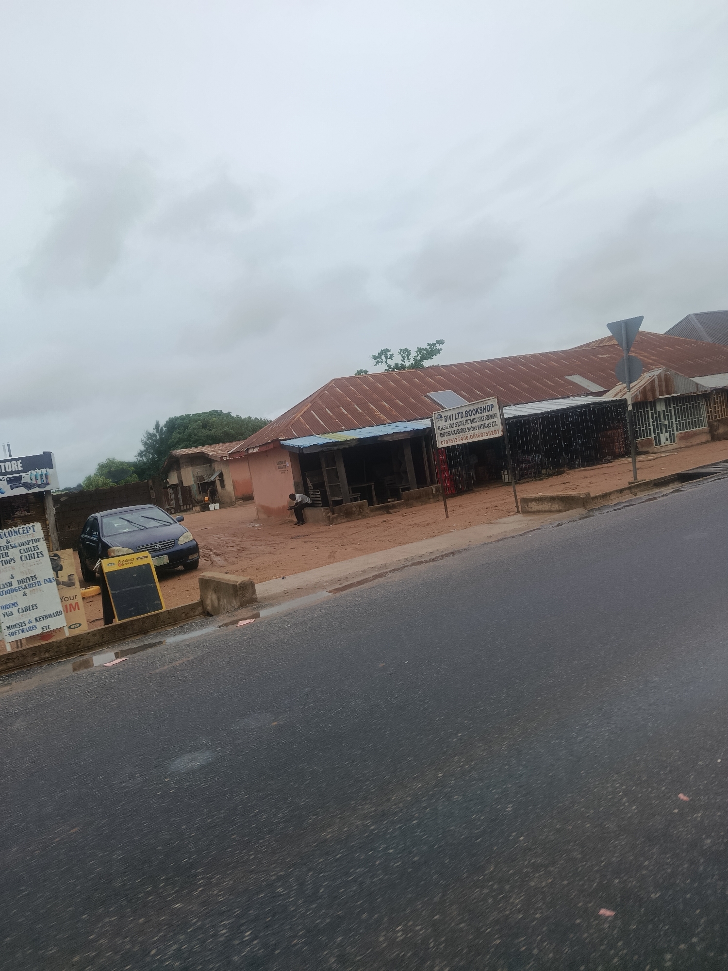 This image captures a street scene in Jalingo, Nigeria, on an overcast day. The foreground features a wet asphalt road with a slight sheen, indicating recent rain. On the right side of the road, a concrete drainage channel is visible.
Adjacent to the road, there is a dirt area where several structures are situated. A dark-colored sedan is parked on the dirt, facing slightly towards the camera. In front of the car, a yellow sign with black lettering stands on a metal easel. To the left of the car, a multi-panel sign with text and images is mounted on a brown brick wall. The text on this sign appears to list various products and services, including computer-related items.
Further back, a building with a rusted corrugated metal roof stretches across the scene. A section of this building has an open front with a dark interior, and a person is seated near the entrance. Another sign, "BIVI LTD. BOOKSHOP," is mounted on a pole in front of this building, listing the types of items sold, such as books, stationery, and office equipment. The buildings appear to be single-story structures with some older architectural elements. Trees and greenery are visible in the background, suggesting a semi-urban or suburban setting. The sky is a uniform grey, contributing to the muted lighting of the scene.
