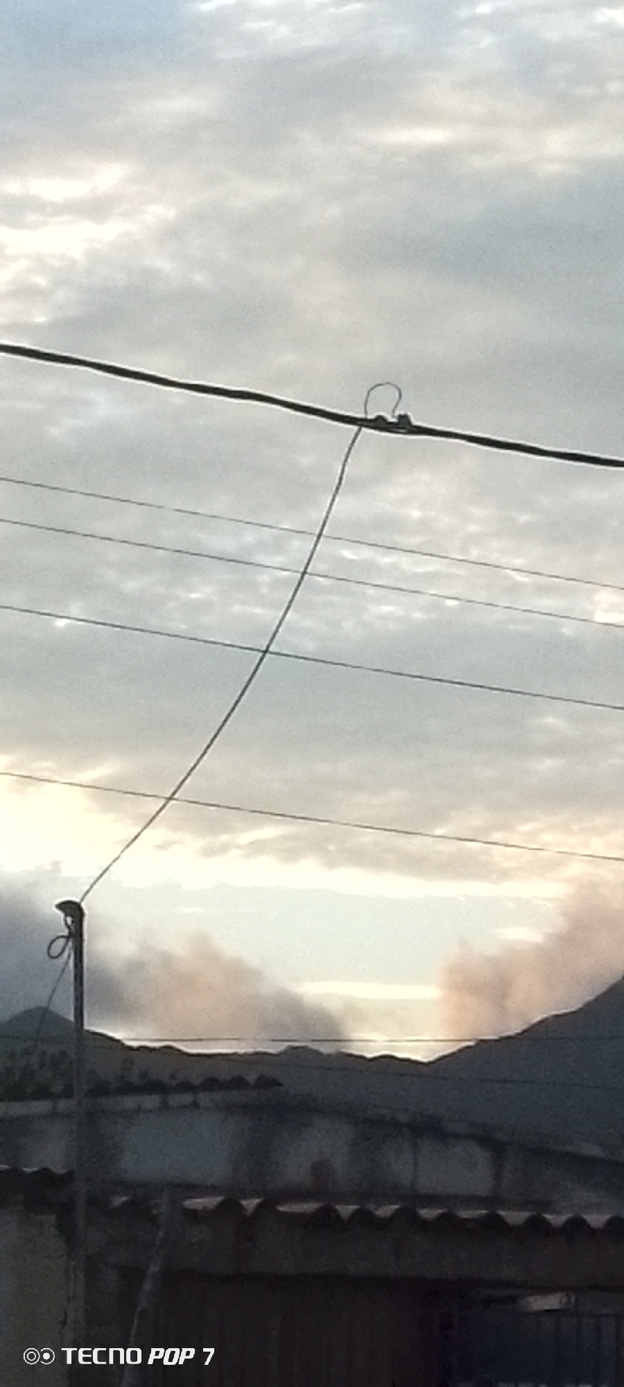 The image shows a cloudy sky over a hilly landscape. In the foreground, there are power lines stretching across the frame. A thin wire dangles from the upper power line, attaching to a pole on the left. The pole itself has a loop of wire around its top. In the background, the silhouette of hills is visible, with some smoke or mist rising from the landscape. The bottom of the image shows the top of a building with a corrugated roof. A watermark in the bottom left corner reads "TECNO POP 7". The scene appears to be outdoors, possibly in a rural or semi-rural area. The lighting suggests it is either early morning or late afternoon.