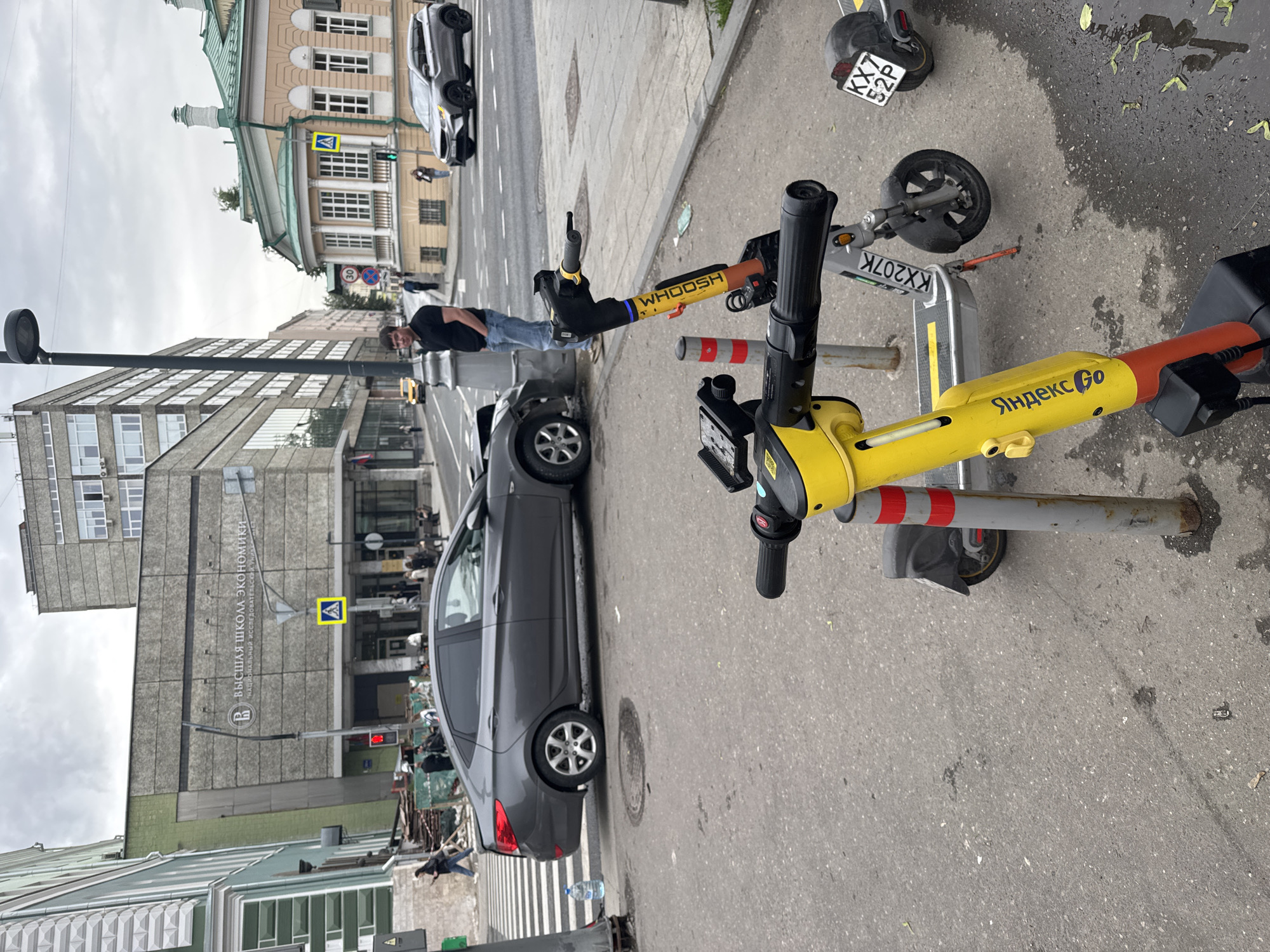 The image shows a Moscow, Russia street scene with parked rental scooters and a damaged car. The car, a gray sedan, is parked at the curb with damage to its front driver's side, near a person lying on the sidewalk. Multiple scooters with the "Yandex Go" and "Whoosh" branding are parked nearby. A person is standing near the damaged car, and other pedestrians are visible further down the sidewalk. Buildings in the background include a high-rise with the inscription "ВЫСШАЯ ШКОЛА ЭКОНОМИКИ" (Higher School of Economics) and another building with a green roof. A 30 km/h speed limit sign is also visible.