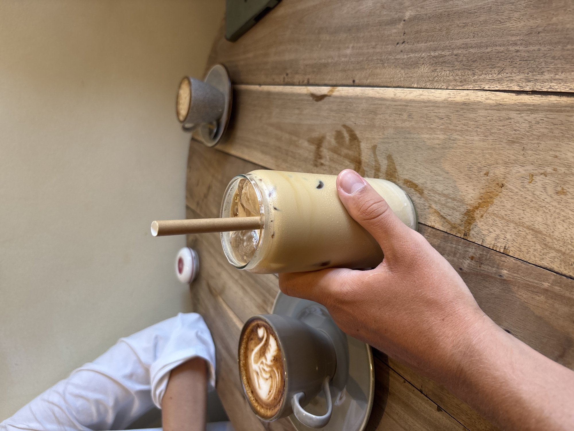 A person's hand is holding a glass of iced coffee with a straw on a wooden table. There are two additional cups of coffee on the table, one containing latte art. A white shirt sleeve is visible in the lower left corner of the image. The scene is likely located in a cafe, judging by the coffee and table setting. The photograph was taken indoors, with natural lighting. This scene takes place in Kuta Selatan, Indonesia.