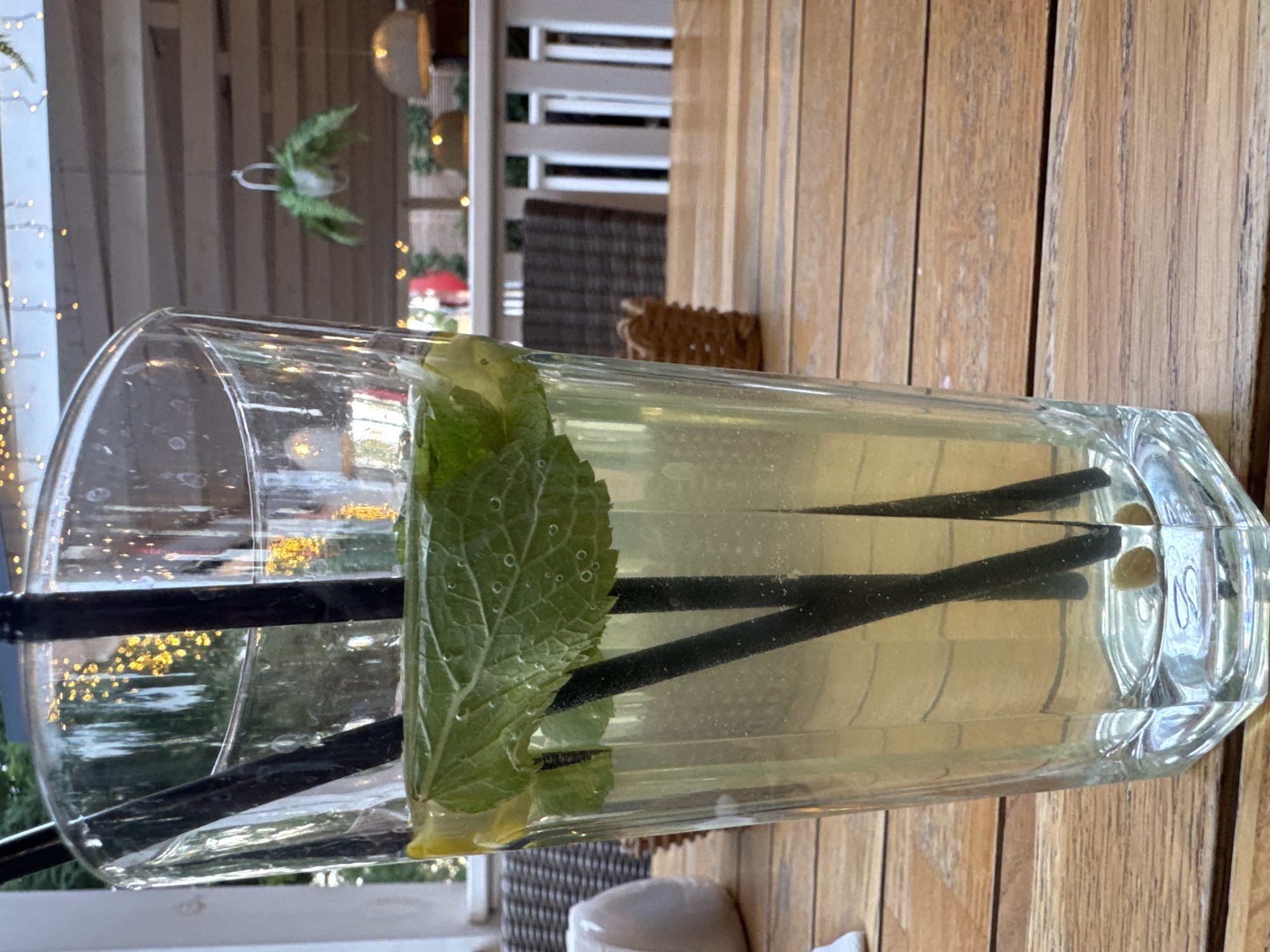 A close-up, eye-level shot shows a tall, clear glass filled with a pale yellow, bubbly liquid and garnished with mint leaves and lemon slices. Two black straws are submerged in the drink. The glass sits on a light brown, wooden table. In the background, out of focus, is a covered patio area with hanging lights and greenery. The scene suggests an outdoor cafe or restaurant setting in Yevpatoriya, Ukraine.