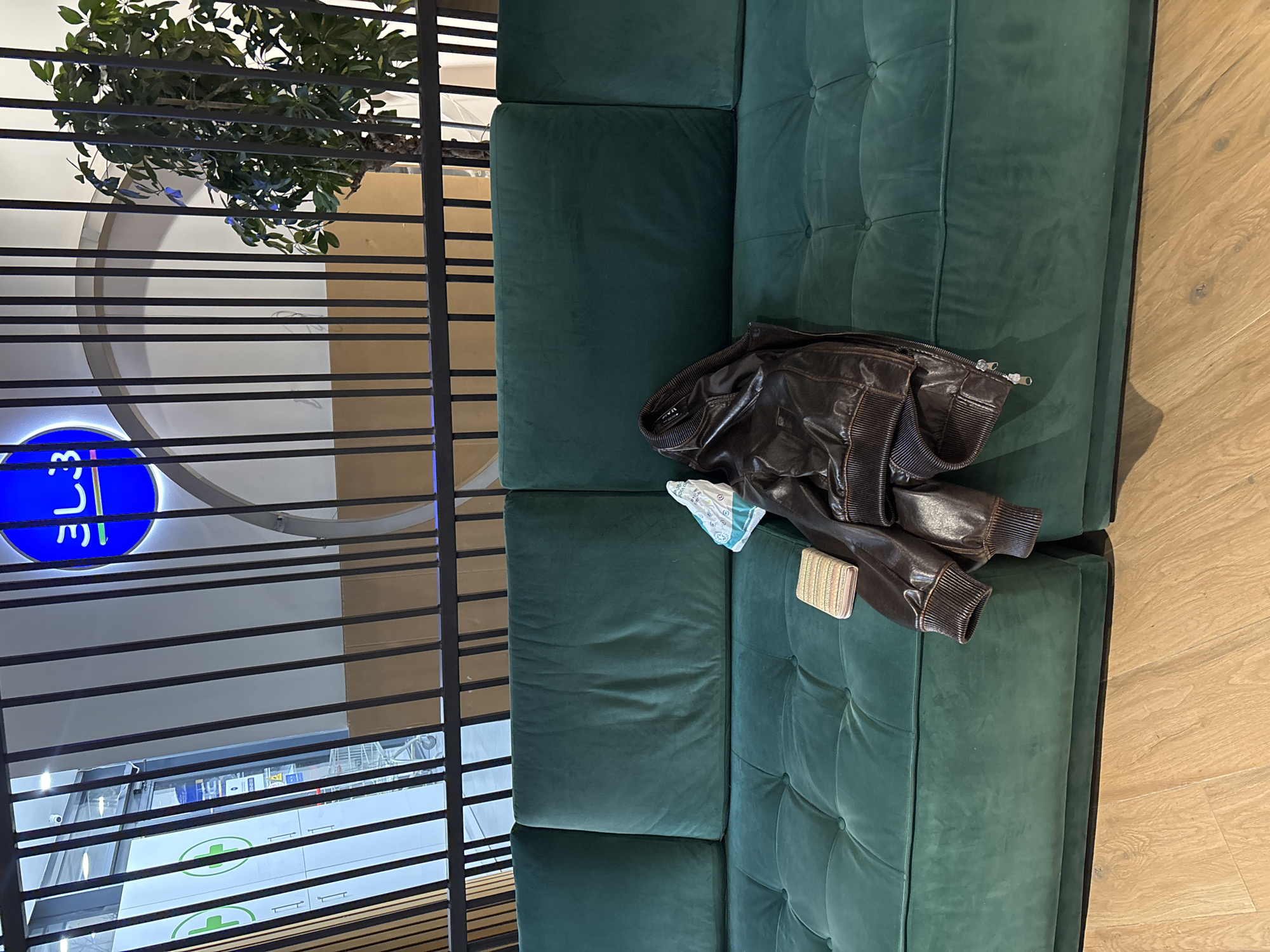 The image shows a section of an indoor waiting area, likely in a commercial or public building. The viewpoint is from slightly above, looking down at a green, tufted couch. A dark brown leather jacket is casually placed on the couch, accompanied by a face mask and a small, metallic clutch or wallet.
In the background, through what appears to be a metal slat screen, portions of the building's interior can be seen. This includes a blue sign with white lettering (possibly an advertisement or store indicator), architectural details, and what appears to be a shop counter or shelving. A potted plant or tree is visible above the screen.
Based on the blue sign and the appearance of what may be a pharmacy counter, the location is potentially in Tbilisi, Georgia, as there are establishments with similar visual elements in that city. However, specific store names cannot be determined with certainty from the image.