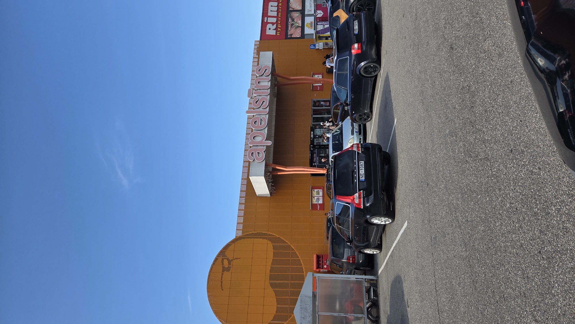 An exterior, eye-level, vertical shot shows the "Apelsins" supermarket in Stapriņi, Latvia, under a clear blue sky with a few wispy clouds. The supermarket building is primarily orange with large, pink lettering that reads "apelsins". To the left, a large, circular orange design is visible on the building's facade. The entrance area is marked by orange poles supporting a white overhang. To the right of the entrance, signage for "Rimi Hypermarket" is present. The foreground is dominated by a black asphalt parking lot with white parking lines. Several cars are parked in the lot, including a black Volvo station wagon in the center with license plate "NE-1087", a silver sedan, a dark blue Audi station wagon with license plate "HB-1010", and other vehicles partially visible. A grey trolley shelter with red elements is situated on the far left of the image. A few pedestrians are visible near the supermarket entrance. The overall scene depicts a typical day at a retail establishment with a parking lot.