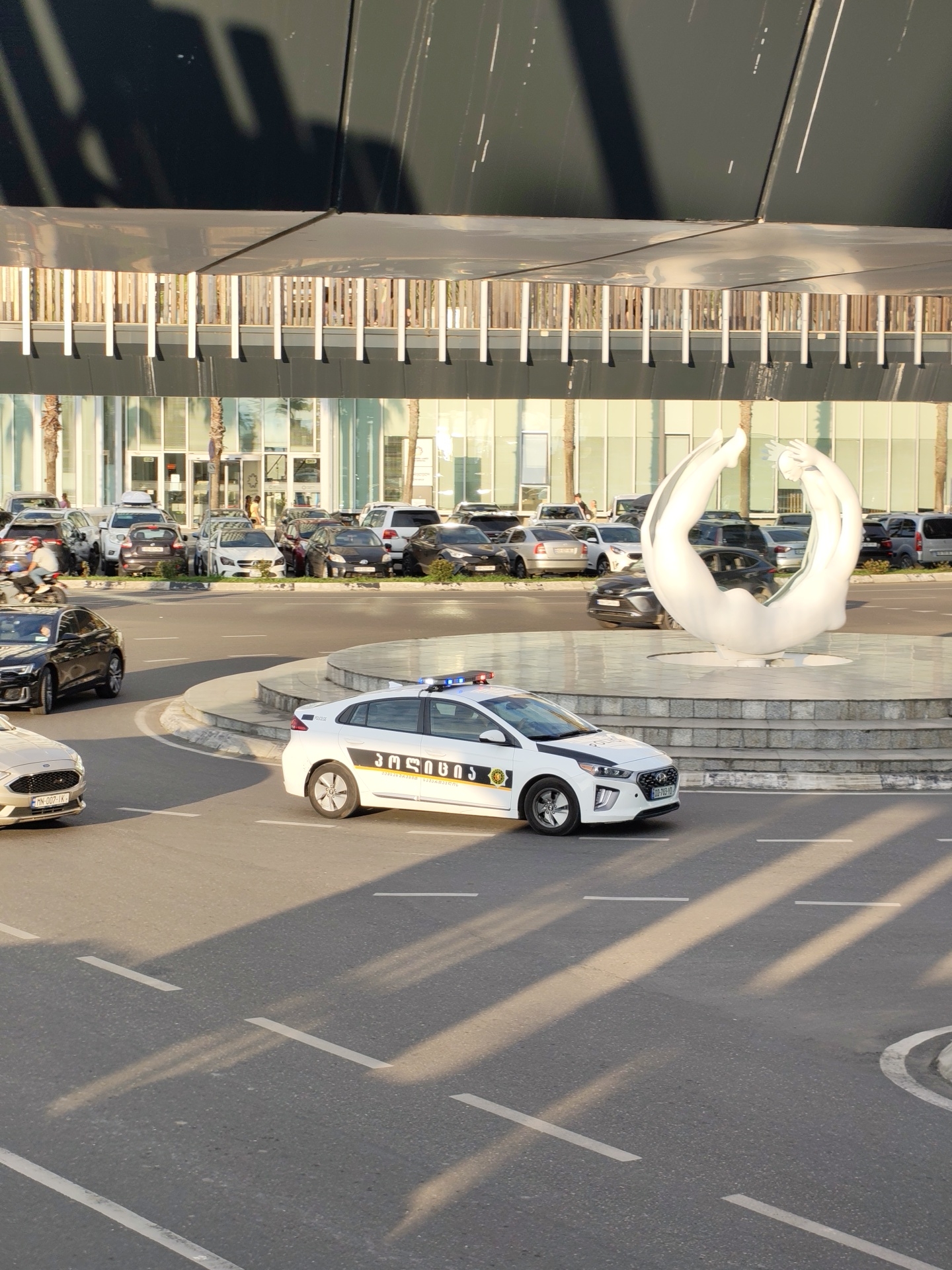This image captures a street scene in Batumi, Georgia, featuring a white police car with flashing blue and red lights. The car has Georgian text on its side and a black stripe across the hood. It is positioned on a paved road with white lane markings, suggesting it is in a traffic area. In the background, a modern building with glass windows and an overhanging structure is visible. Adjacent to the road, a circular fountain area with a large, abstract white sculpture of intertwined figures stands. Several parked cars are visible behind the fountain, and a few palm trees are scattered throughout the scene. The lighting indicates it is daytime, with shadows cast by the overhanging structure and the vehicles.