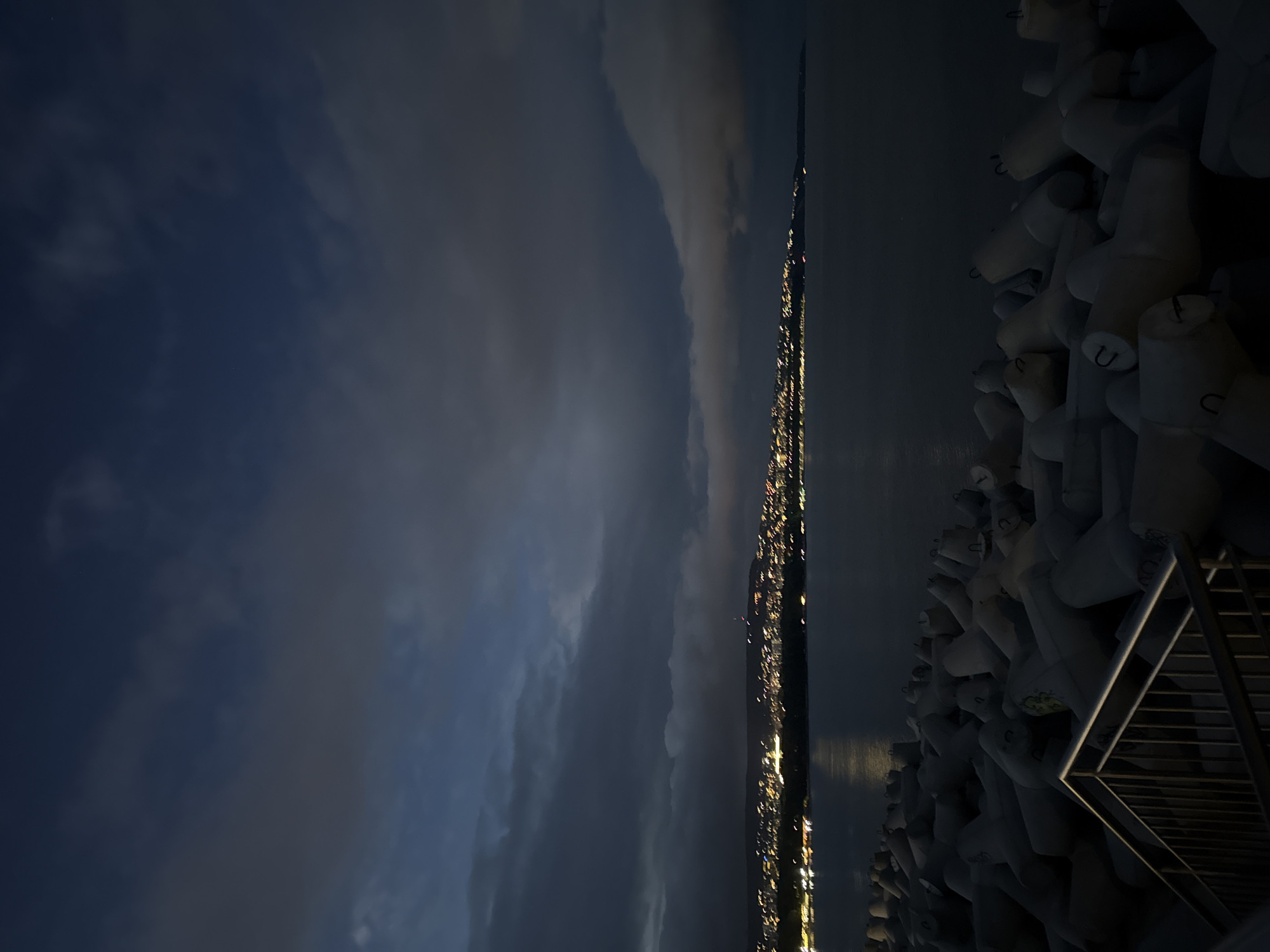 The image captures a nighttime scene in an unknown city. The sky is filled with dark, heavy clouds, with subtle gradations of blue. A thin, illuminated cityscape stretches across the horizon. The lights suggest a dense urban area. Adjacent to the city, concrete structures, possibly tetrapods or other coastal defenses, are densely packed together. A metal railing and stairs are visible, suggesting a public access point to the coastal area.