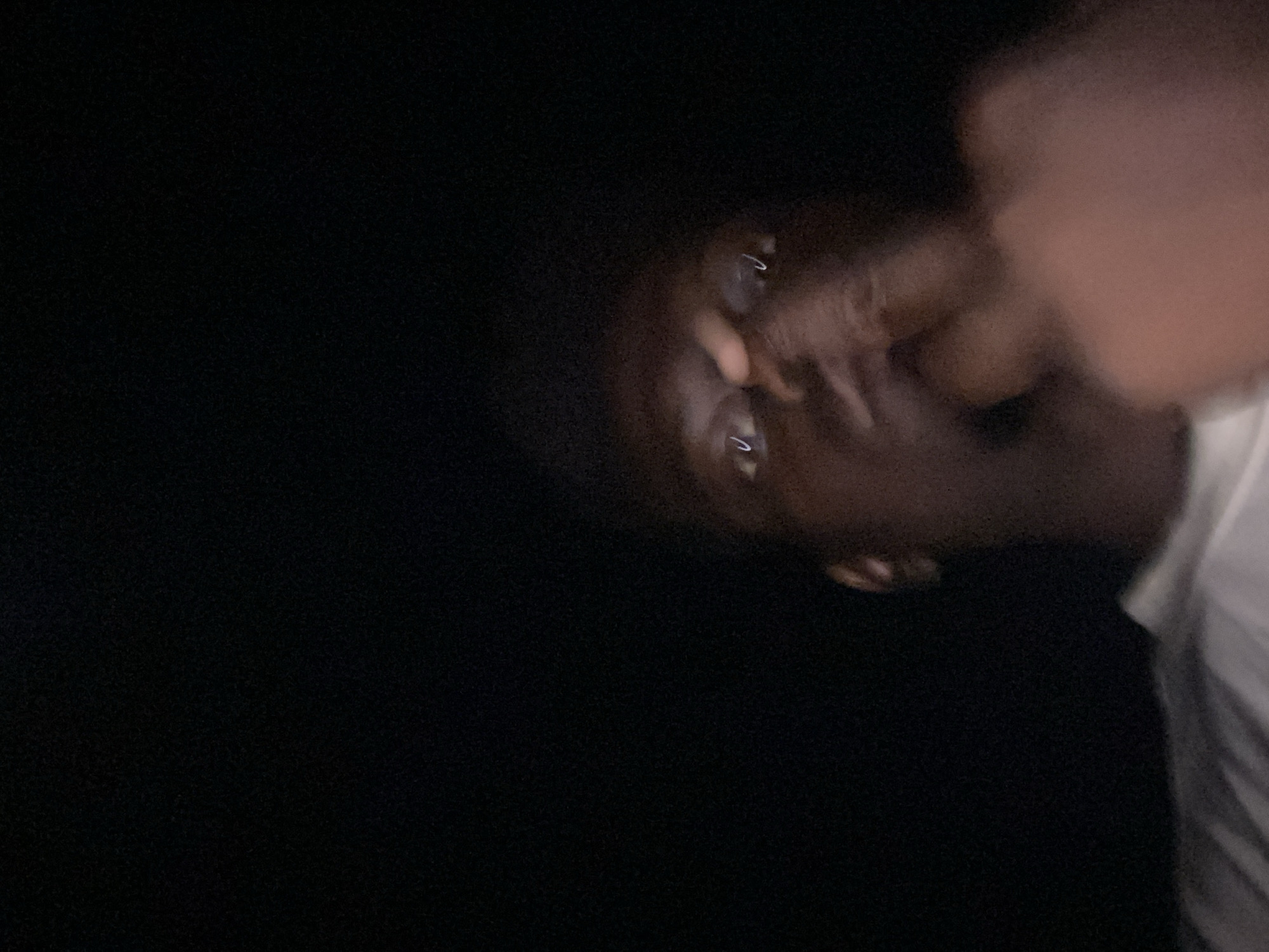 The image presents a close-up, diagonally oriented view of a person's face against an entirely black background. The subject, who has dark skin, is positioned on the right side of the frame, appearing to look towards the viewer or camera. One eye is clearly visible with a distinct 'C' or 'U' shaped reflection, and a second eye is partially seen below it. The bridge of the nose is discernible.
A blurry hand or fingers appear to be in close proximity to the person's face, possibly touching or partially obscuring the mouth or cheek area. On the far right edge of the image, a white fabric, likely part of a shirt, is visible, indicating the presence of clothing.
The scene is set in complete darkness, strongly suggesting that the photograph was taken at night or in an unlit indoor environment. This lack of ambient light contributes to the overall blurriness of the subject's face and features. Due to the low light and blur, the person's emotional state is not discernible. There are no other objects or specific setting details visible, nor is there any visible text.