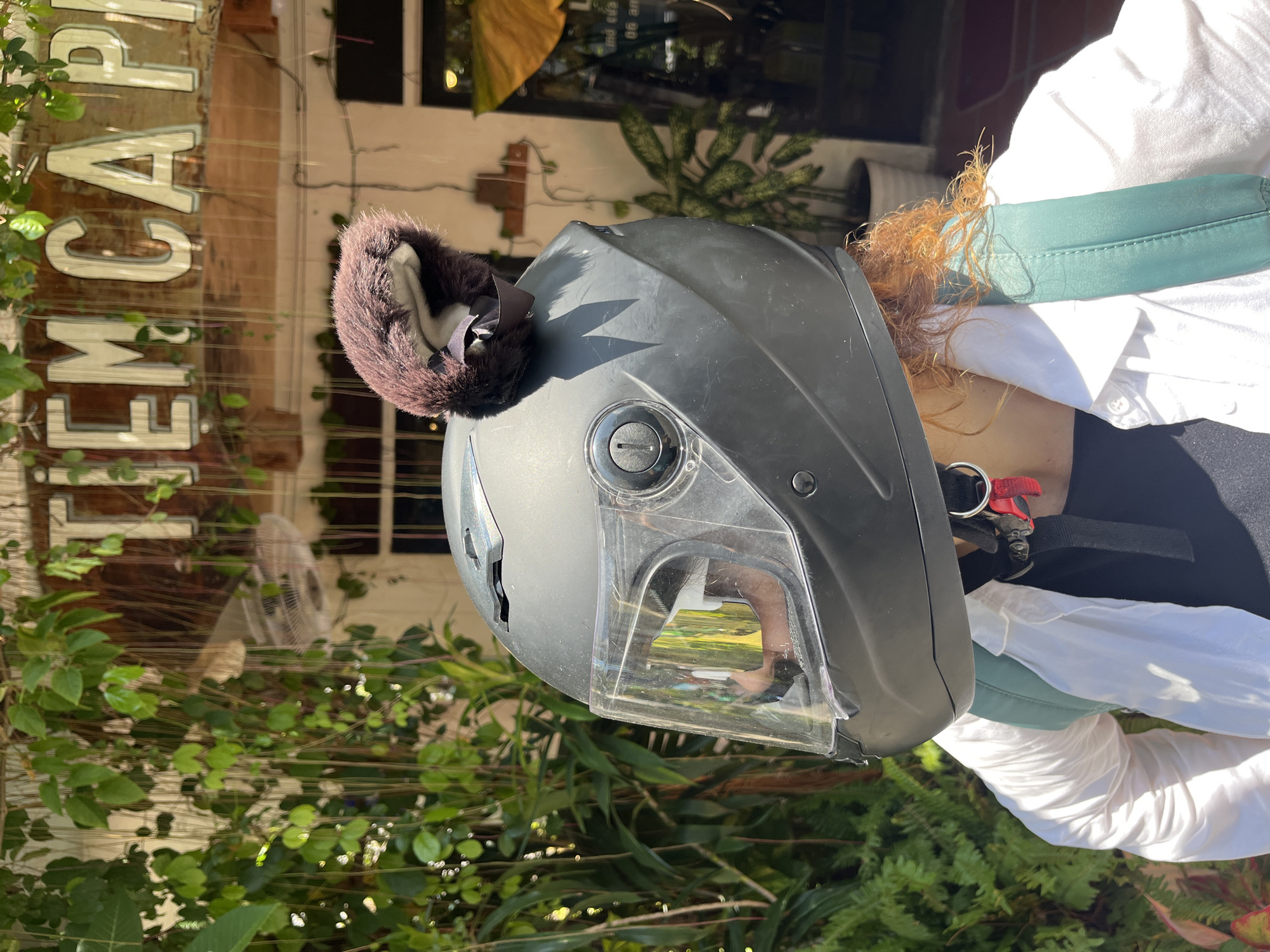 A person wearing a black motorcycle helmet with fuzzy brown ear attachments and a white shirt stands in front of a building with a sign that reads "TIEM CA PH..." and lush green foliage. The helmet has a clear visor, reflecting the surroundings. The person also appears to be wearing a green backpack. The scene is outdoors, with bright sunlight.