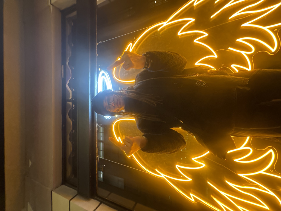 A person wearing a dark outfit, glasses, and a hat poses in front of a window with a neon wing sign. The person is giving the middle finger with both hands, pointing towards the wings. The sign is a glowing yellow color. The window frame is visible, along with hints of the building's exterior and the reflected interior. The lighting is dim and emphasizes the bright neon sign.