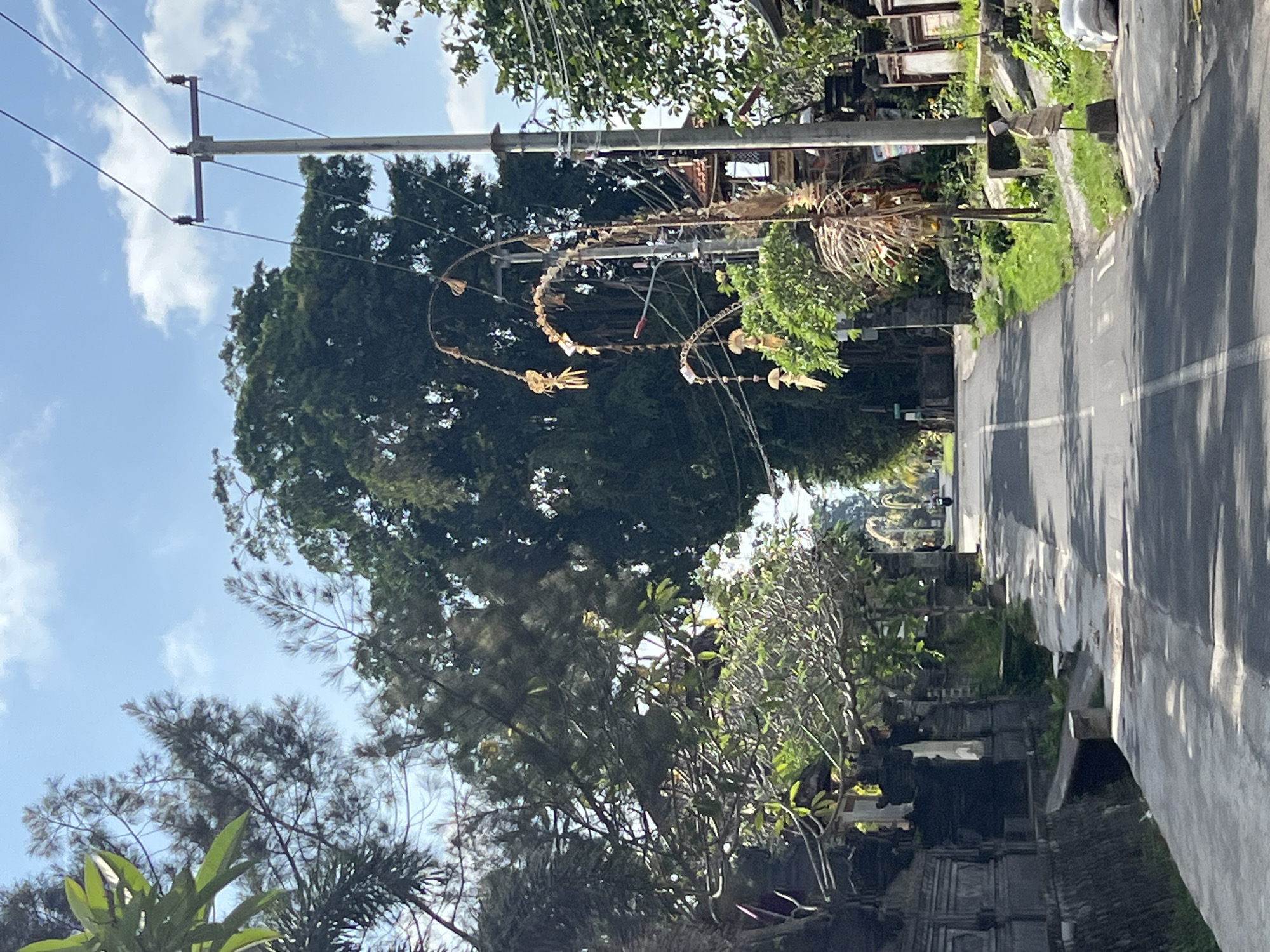 This image captures a street scene in Sukawati, Indonesia. The road, paved with asphalt and marked with a white center line, stretches into the distance, lined with traditional Balinese architecture on the left and lush greenery on the right. Elaborate, traditional Balinese decorations, likely Penjor, are prominently displayed along the roadside, featuring intricate woven elements and hanging ornaments. A utility pole with multiple wires stands on the right side of the road, indicating the presence of infrastructure. In the background, a lone individual on a motorcycle is visible, suggesting the road is in use. The scene is bathed in bright sunlight, casting shadows and highlighting the textures of the stone walls and natural foliage. The sky above is a clear blue with scattered white clouds.