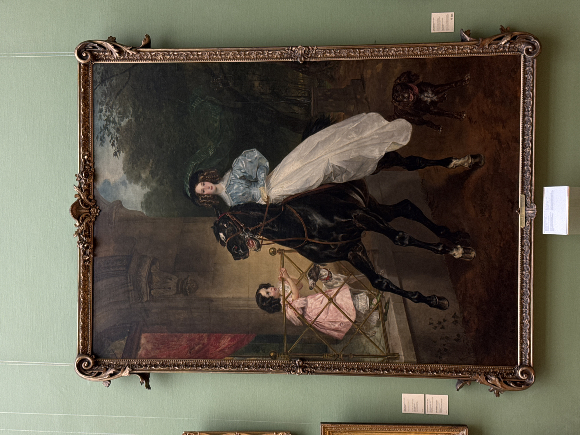 The image shows a framed oil painting hanging on a green wall. The painting depicts a woman in a blue and white dress riding a black horse. Below her, a young girl in a pink dress is inside a metal cage with a white dog. A brown dog is depicted in the background. The style of the painting suggests a historical period. There are informational cards underneath the painting and to the right. The setting is likely a gallery or museum in Moscow, Russia.