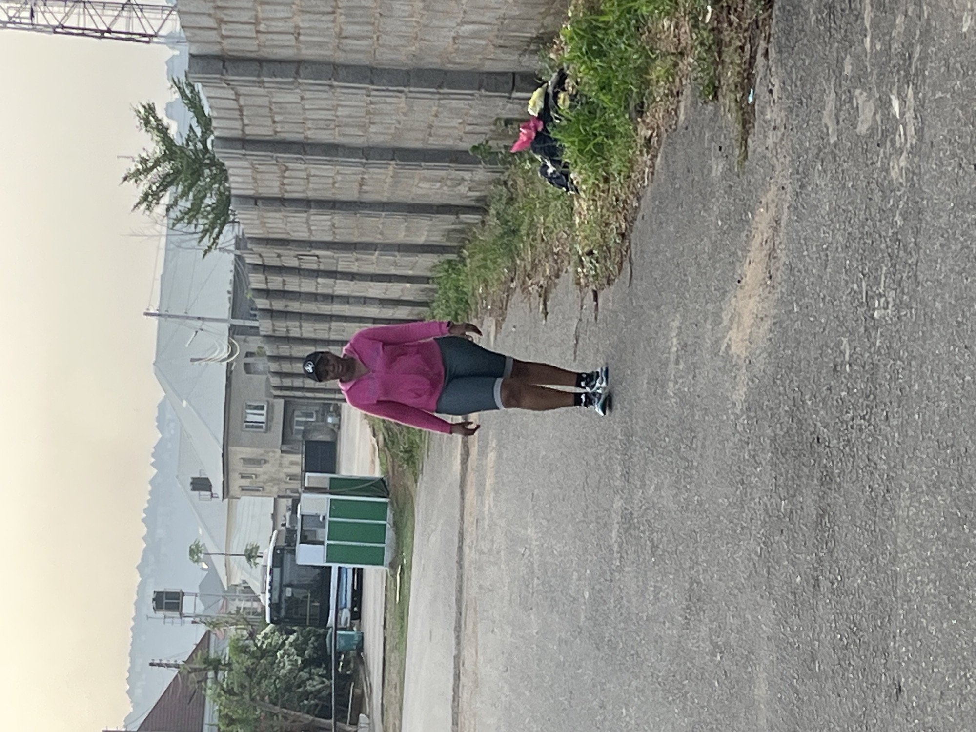 A woman in a pink long-sleeved shirt, gray shorts, a black hat, and black and white athletic shoes is standing on a gray asphalt road. The road is lined on the left with a concrete block wall and on the right with overgrown grass. Behind the woman are a few buildings, trees, and a range of hills in the distance. A green and white rectangular structure is also visible near the buildings. This appears to be a residential area in Abuja, Nigeria.