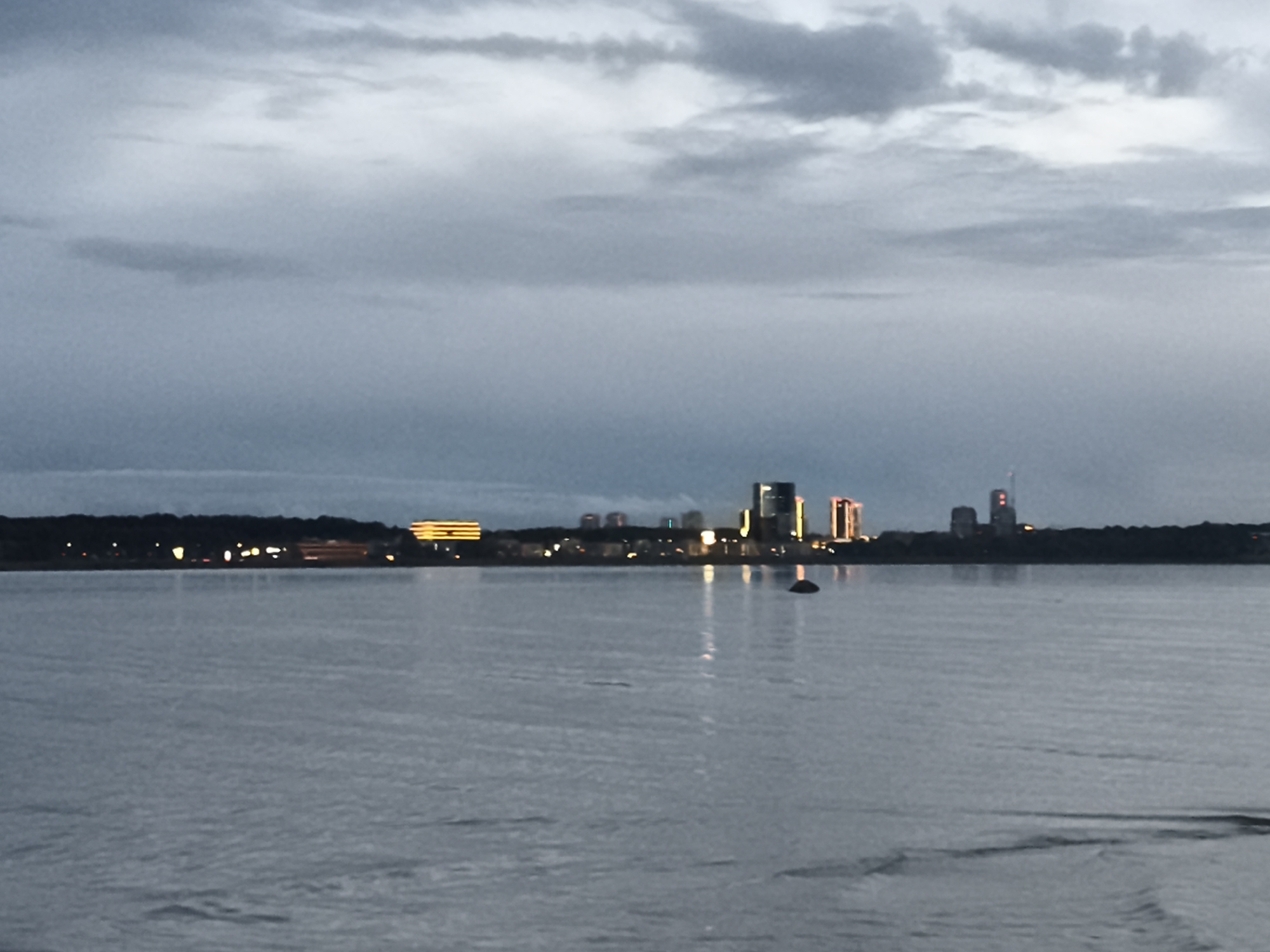 The image captures a serene twilight view of the Tallinn skyline across the water, likely the Tallinn Bay. The water in the foreground is calm, with subtle ripples reflecting the muted light. The distant shoreline is marked by a dark tree line, beyond which a cluster of illuminated buildings stands out. Among these, a prominent structure with horizontal yellow lighting is visible on the left, resembling a hotel or office building. To its right, several modern high-rise buildings with illuminated windows and some colored lights, possibly red and white, form the central part of the city silhouette. A solitary dark, rounded object, possibly a buoy or a small vessel, is present in the water to the right of the illuminated buildings. The sky above is overcast with grey clouds, creating a soft, diffused light typical of dusk or dawn. The overall impression is one of a peaceful urban panorama at the edge of the sea.