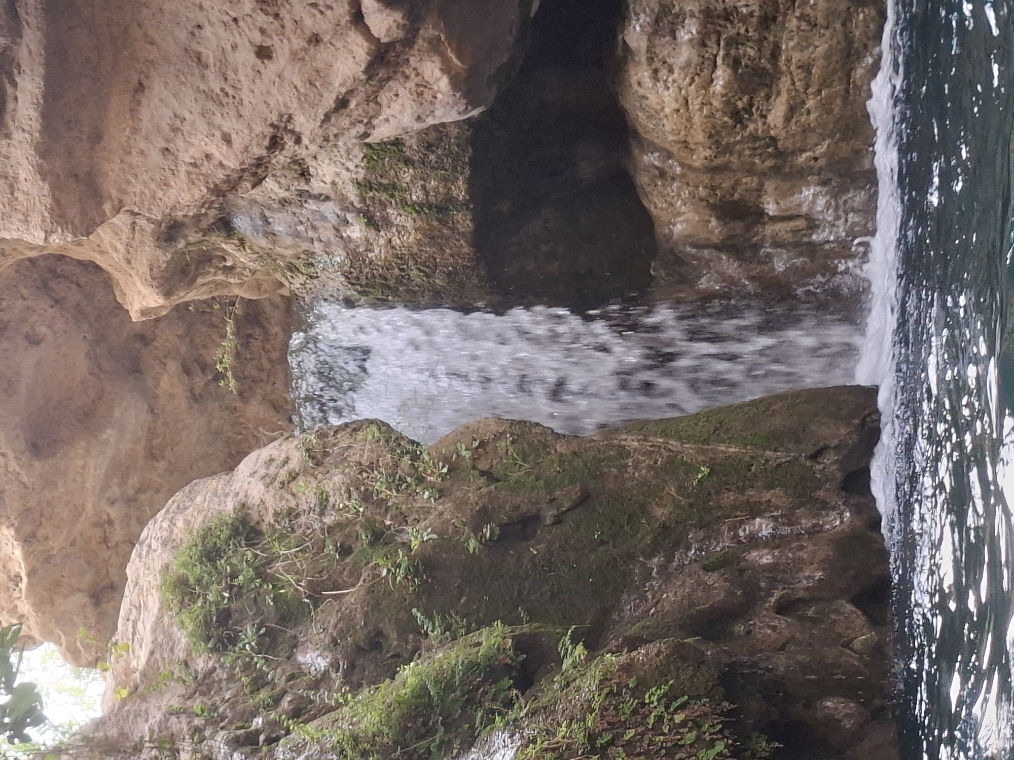 A vertical shot captures a narrow waterfall cascading between rocky canyon walls. The water, a frothy white, plunges into a dark pool below, creating ripples and splashes on the surface. The canyon walls are composed of rough, weathered rock in shades of tan and brown, with patches of moss and small green plants clinging to their surfaces. The scene is dimly lit, suggesting it's within a shaded gorge.