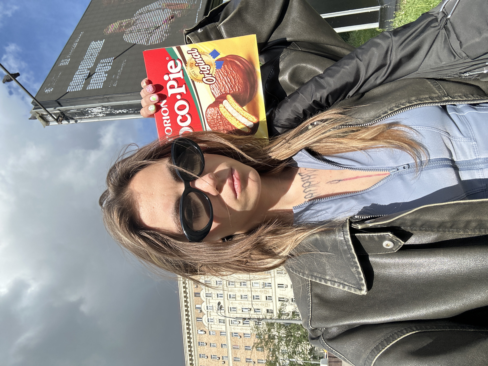 A medium shot captures a young woman holding a box of Orion Choco Pie in Moscow, Russia. The woman is wearing black sunglasses, a blue zippered top, and a dark leather jacket. She has blonde hair and visible tattoos on her chest. In the background, a large building with many windows, a street lamp, and a billboard can be seen. The sky is partly cloudy.