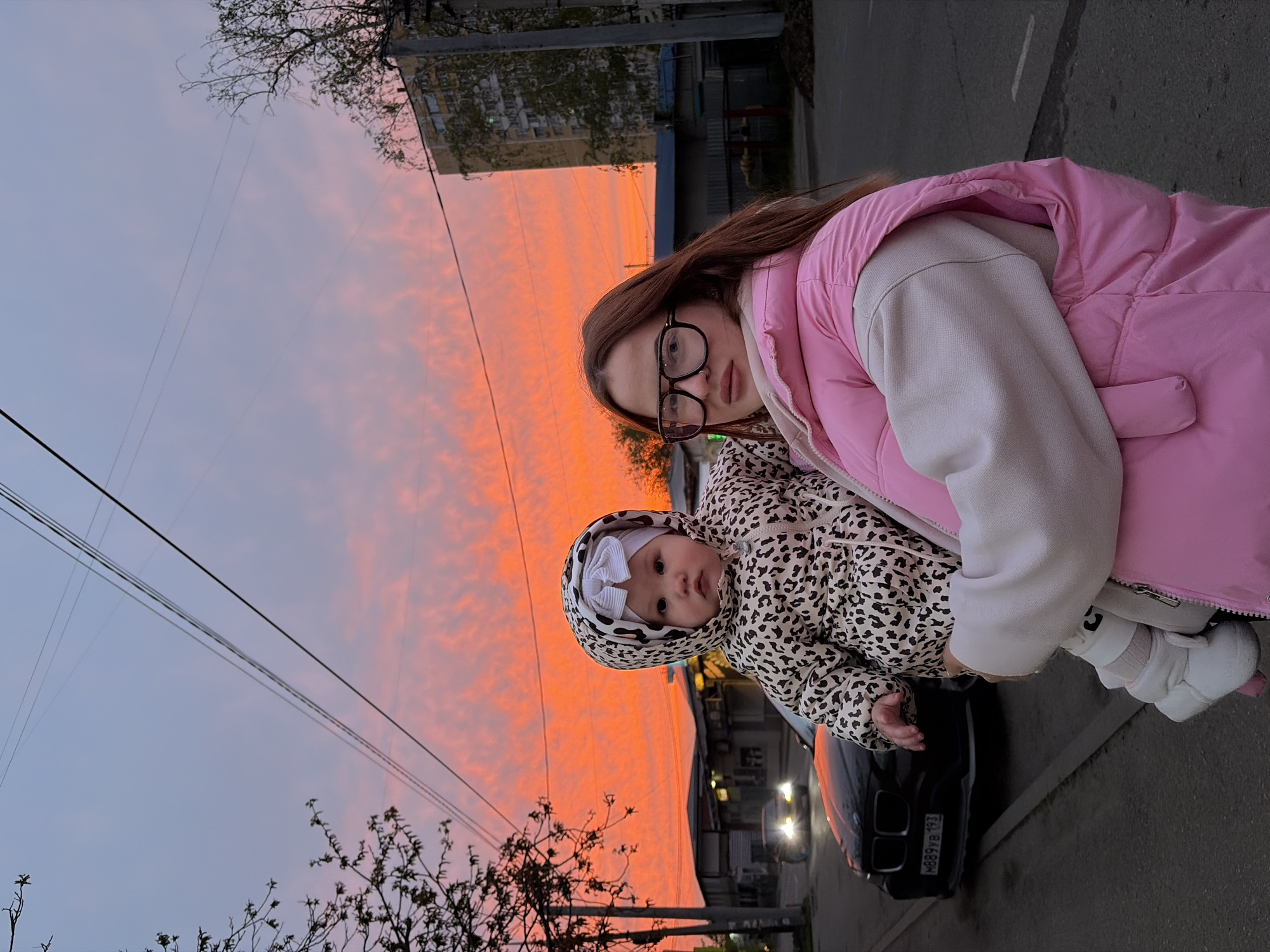 The image, appearing rotated 90 degrees counter-clockwise, depicts a young woman holding an infant outdoors at sunset in Krasnodar, Russia.
The woman, positioned on the right (when viewed upright), has long brown hair and wears black-rimmed glasses, a light-colored top, and a prominent bright pink puffer vest. She looks calmly towards the camera. She holds a baby, dressed in a leopard print hooded jacket and a white bow headband, who gazes curiously slightly off-camera. Both individuals are dressed in attire suitable for cool weather.
The background is dominated by a dramatic sunset sky, transitioning from a soft blue and pale pink at the top to vibrant orange and red hues closer to the horizon, indicative of late evening. Several power lines crisscross the sky. To the left, bare tree branches are silhouetted against the colorful backdrop. In the midground, a black car is parked on an asphalt street; its rear lights are dimly lit, and a license plate, "M889YB 93," is visible, confirming the Russian Krasnodar region. Buildings can be seen in the distance beneath the horizon. The scene captures a tranquil moment against a striking natural light display.