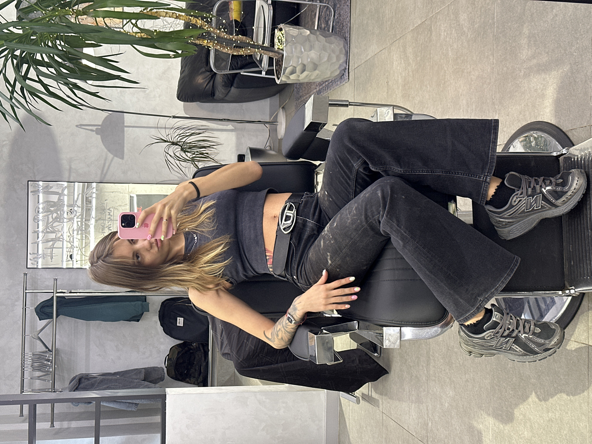 A young Caucasian woman with long blonde hair is sitting in a barber chair and taking a selfie with her pink smartphone. She is wearing a dark grey tank top, dark wash flared jeans, and grey New Balance sneakers. The woman has tattoos on her left arm and a black bracelet on her right wrist. She is wearing a silver necklace and a belt with a silver buckle. The background appears to be a modern salon or barbershop with grey walls, a clothing rack with jackets, and a large potted plant. The overall lighting suggests it is daytime. The specific location in Moscow, Russia is not determinable from the image.