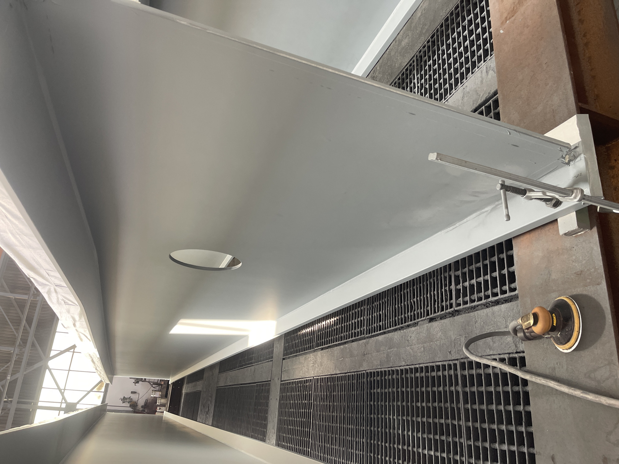 The image shows a large, flat, gray metal structure suspended above a grated floor. A circular hole is visible in the metal structure. A sanding tool is attached to the grate. The setting appears to be an industrial environment.