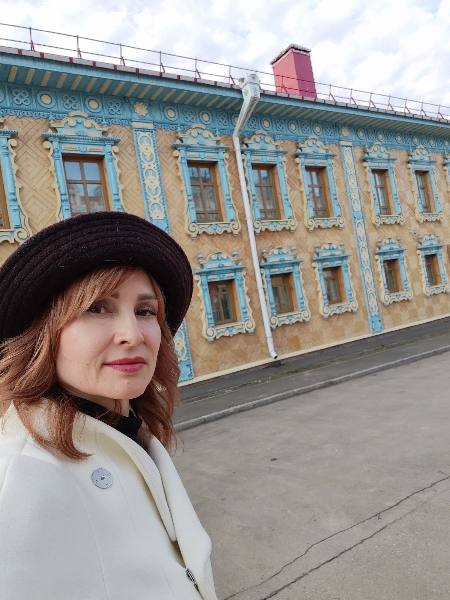 A woman in a brown hat and white coat stands in front of a building with ornate, traditional Russian architectural details. The building has a tan facade with numerous windows framed in light blue, detailed carvings. A red chimney is visible on the roof. The woman is facing the camera, and the photo appears to be a selfie. The location is Sarapul, Russia, as evidenced by the distinctive architectural style of the building in the background. The ground is paved, suggesting an urban environment.