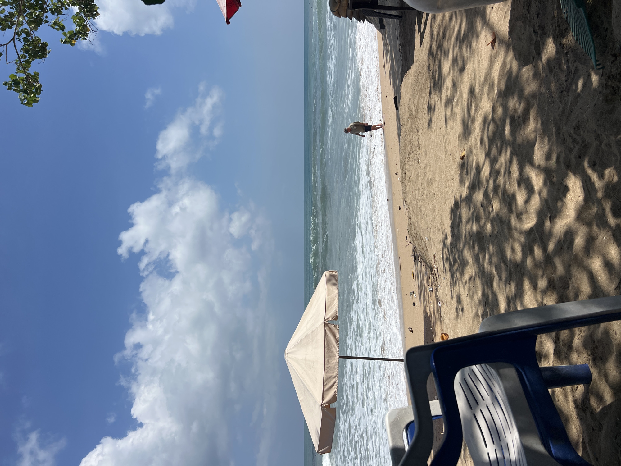 In Kuta, Indonesia, a sandy beach is depicted under a bright blue sky with scattered white clouds. The turquoise ocean features moderate waves breaking near the shore. A light-colored beach umbrella stands prominently on the sand, and in the distance, a lone individual walks near the water's edge. In the foreground, two plastic beach chairs, one blue and gray, and another mostly out of frame, are positioned on the sand. The sand is littered with debris and cast in dappled shade from overhead foliage.