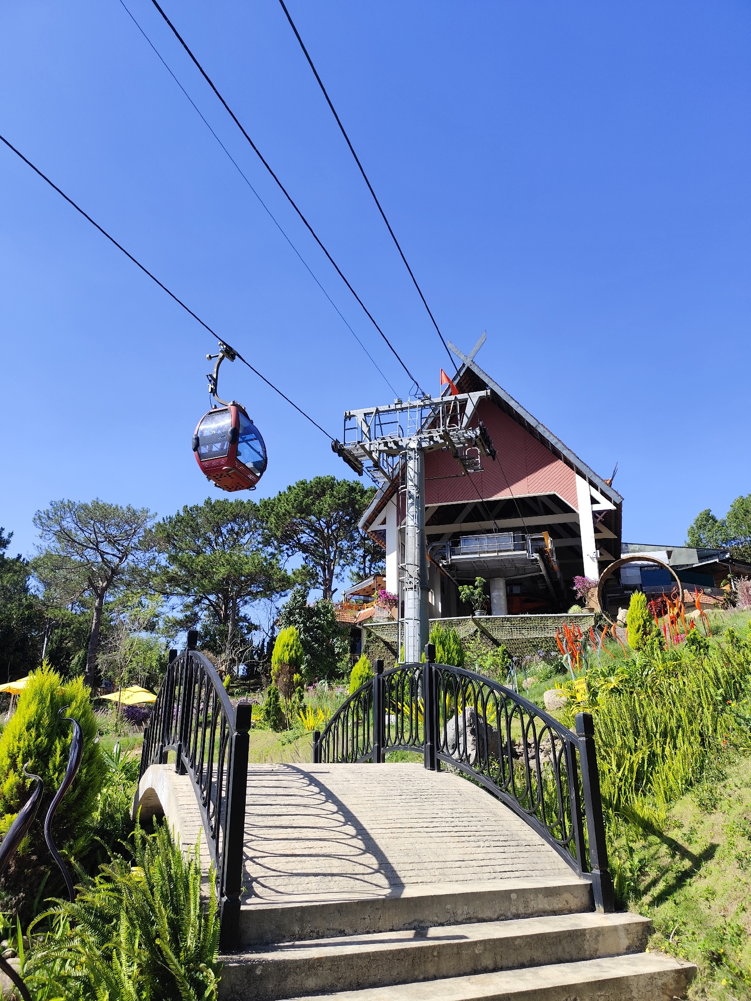 A daytime, eye-level shot shows a cable car cabin approaching a station on a sunny day in Da Lat, Vietnam. A red cable car cabin with blue-tinted windows hangs from overhead wires to the left of a cable car station. The station is red and white and features a traditional Vietnamese roof. A small Vietnamese flag flies on the roof. A second cable car cabin sits in the station on the right. Below the station, there is a lush garden with trees, plants, and flowers. A small concrete bridge with black metal railings is in the foreground, leading up to the station. The sky is blue.