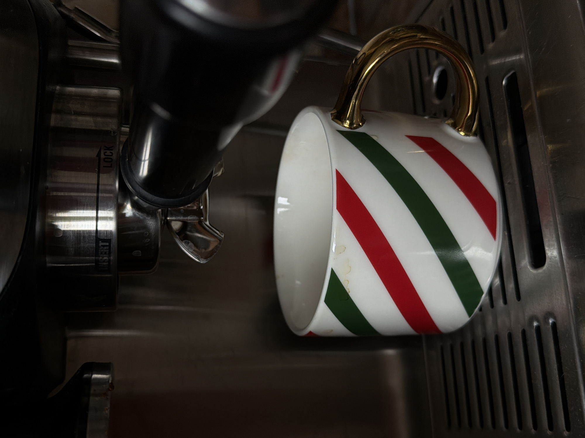 The image shows a white mug with diagonal red and green stripes and a gold handle. The mug is positioned beneath a metal spigot of an espresso machine. There are some stains visible on the mug. The background includes parts of the espresso machine and a stainless steel surface, possibly a sink or drain area. There is no evidence to suggest the location is in Khimki, Russia. The image is focused on the mug and the coffee making process.