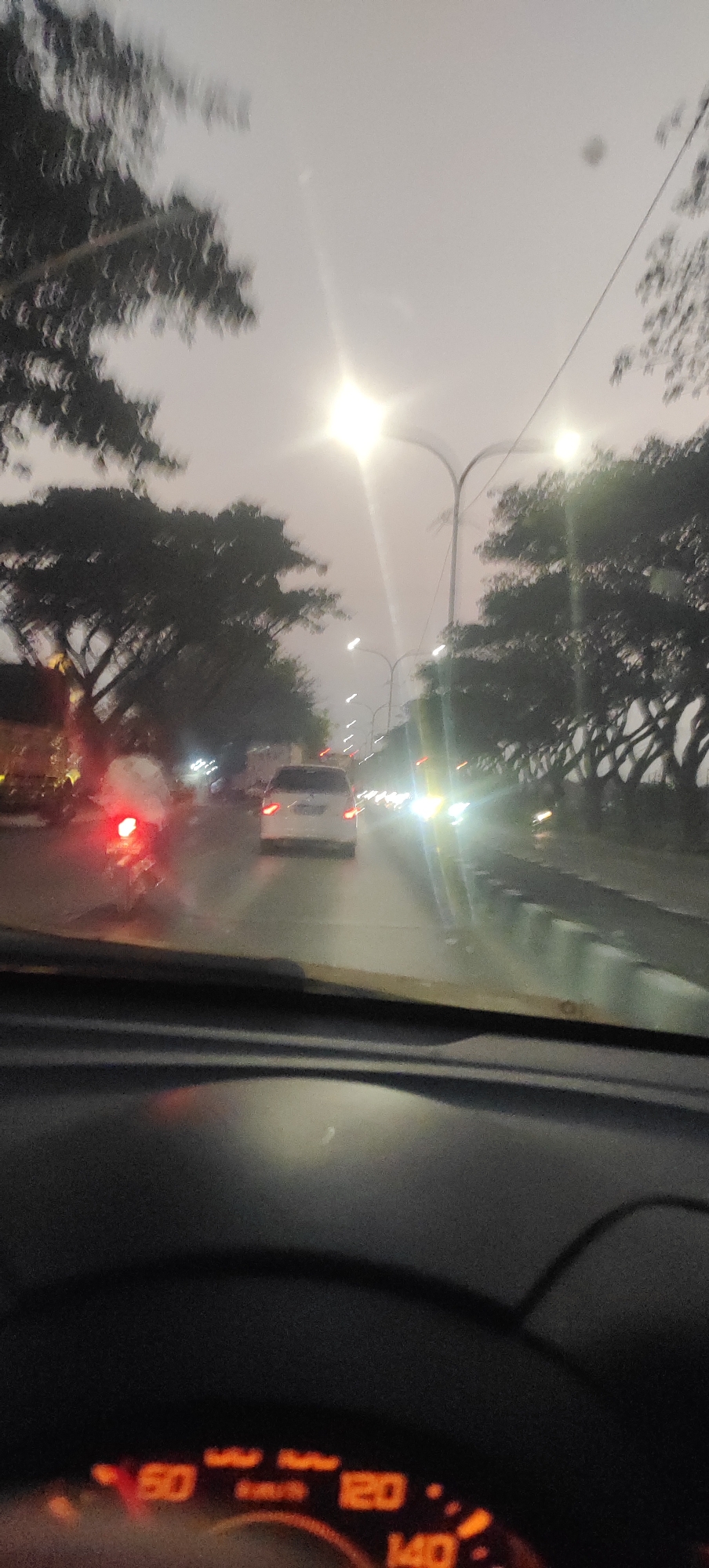 The image is a blurred, overexposed photograph taken from inside a vehicle, likely a car, on a road in Ngemplik Wetan, Indonesia, at dusk or during a hazy day. The viewpoint is from the driver's seat, with the dashboard and a portion of the steering wheel visible in the foreground. The dashboard features an illuminated speedometer with markings that include "50," "120," and "140" in orange digits, suggesting the vehicle is in motion.
The road ahead is wet, reflecting the lights of other vehicles and streetlights. Several cars are visible, with a white car directly ahead. A motorcycle with its red brake light illuminated is to the left of the white car. The road is lined with trees on both sides, their foliage dark and somewhat indistinct due to the motion blur and low light conditions. Overhead, streetlights are present, one of which is particularly bright and appears as a starburst due to overexposure and the camera's lens. The sky is a pale gray, contributing to the overall hazy and dim atmosphere. The presence of the speedometer and the view of traffic indicate the vehicle is actively driving on a public road.