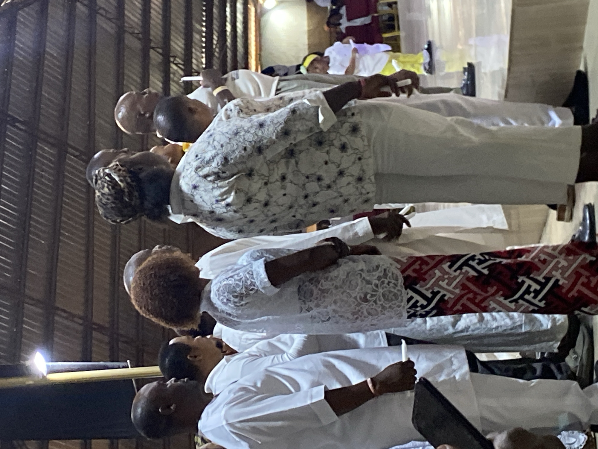 A group of people stand together holding candles in a building with a metal roof. They are wearing white clothing, and the woman in the center is wearing a white lace top with a red and black patterned skirt. The scene is in Rukuba Road, Nigeria.