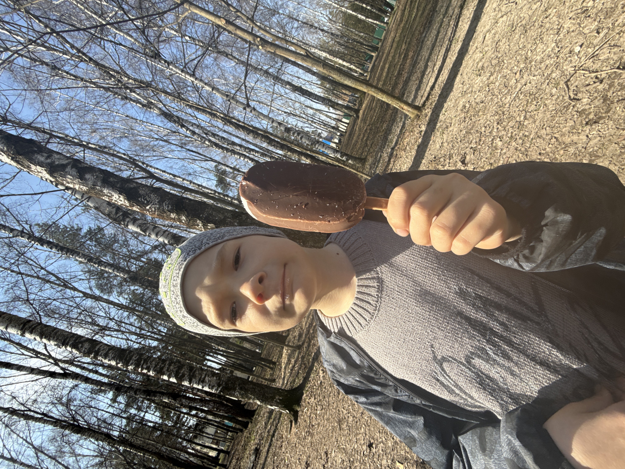A child holds a chocolate-covered ice cream bar in front of them in a park-like setting. The child wears a grey knit hat, a grey sweater, and a dark jacket. The background consists of a grove of bare trees against a blue sky, typical of early spring. The ground is covered in dry leaves. This could be a park or recreational area within Dmitrov, Russia, but without additional information, specific localization is impossible.
