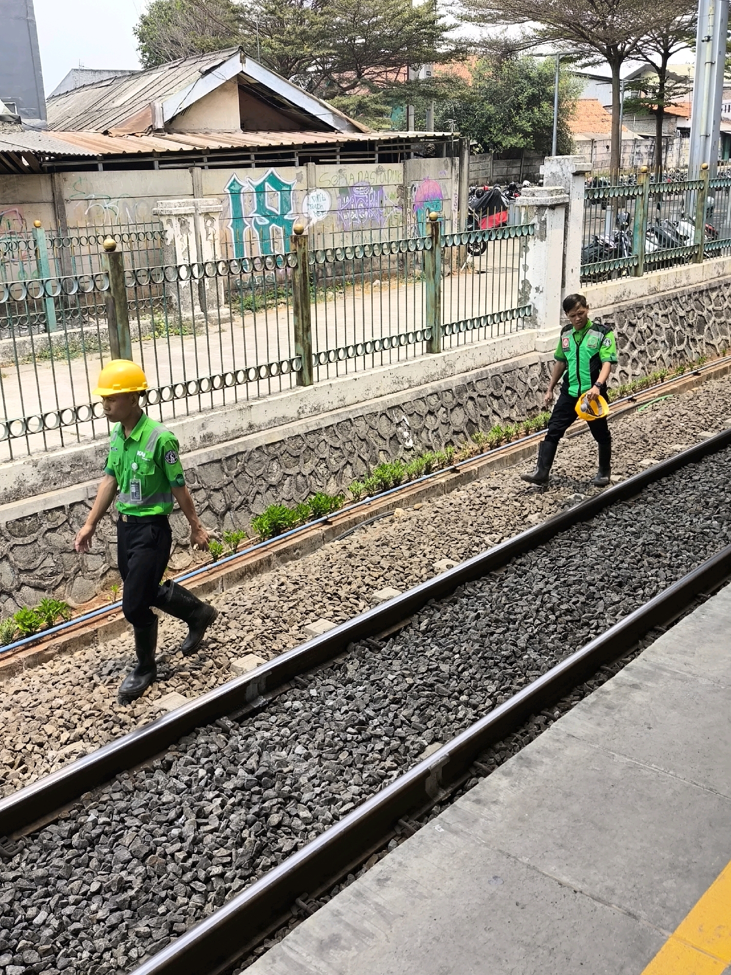 Two male workers in green uniforms and black pants are walking on a railway track in an urban environment. The worker on the left is wearing a yellow hard hat and black boots. The worker on the right is carrying a yellow hard hat and is also wearing black boots. They are walking away from the camera, on separate tracks. The background shows a concrete wall with graffiti, a metal fence, some trees, and buildings. The scene appears to be in Mekarsari, Indonesia, likely near a railway station or maintenance area.