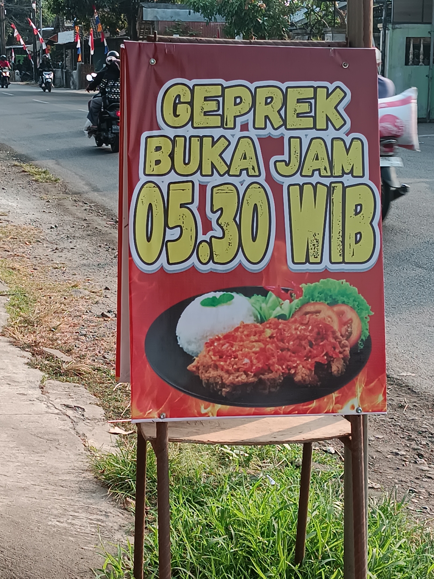 A red banner advertising "GEPREK BUKA JAM 05.30 WIB" is displayed on a metal stand on the side of a street in Imogiri, Indonesia. The banner features a picture of fried chicken with chili sauce, rice, and vegetables. The background shows a road with several motorcycles and people. The street is lined with grass and concrete.