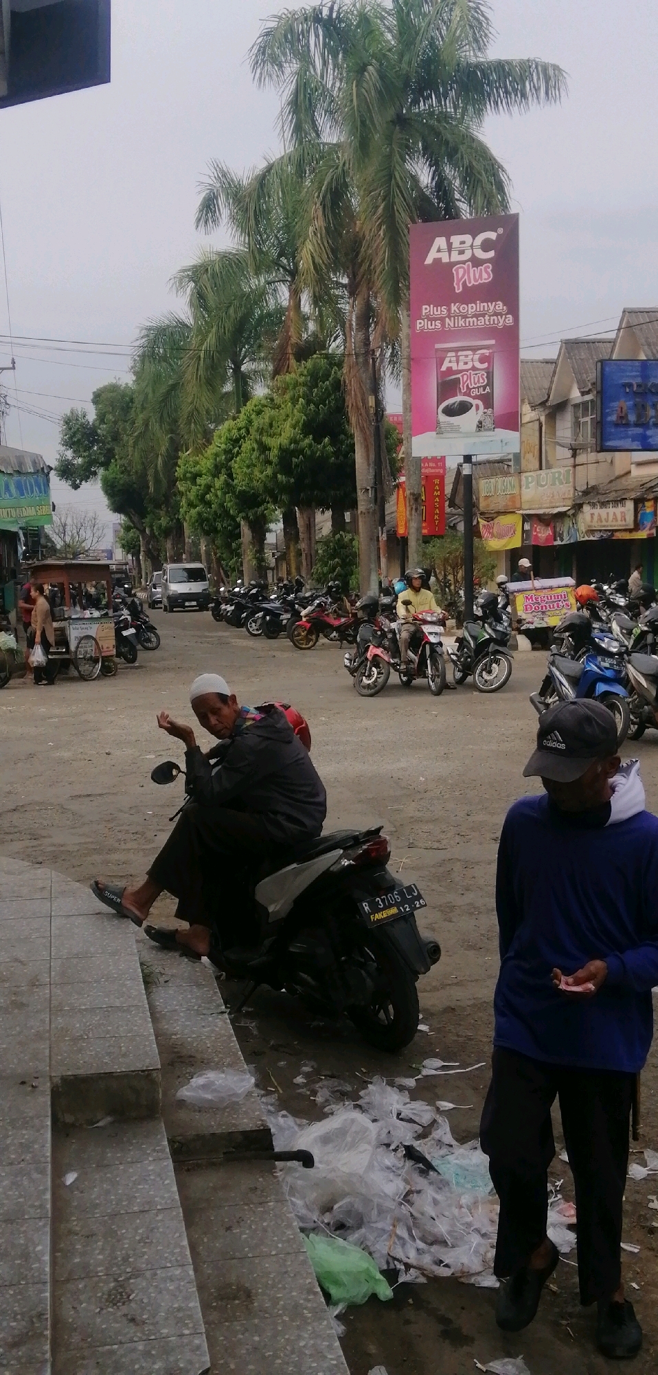 The image captures a street scene in Ajibarang, Indonesia, likely in front of shops and palm trees. In the foreground, a man wearing a white cap and a blue long-sleeved shirt stands, holding some small pink bills in his right hand. To his left and slightly behind, another man is seated on a dark motorcycle, wearing a white skullcap, a dark jacket, and dark pants. He is gesturing with his right hand. The ground is a dirt and concrete mixture, with discarded plastic bags and trash piled up on the right side of the image. Several other motorcycles are parked in the mid-ground, and a street vendor's cart is visible on the left. A large billboard advertising "ABC Plus" coffee is prominently displayed on a pole, partially obscured by palm trees. The sky is overcast.