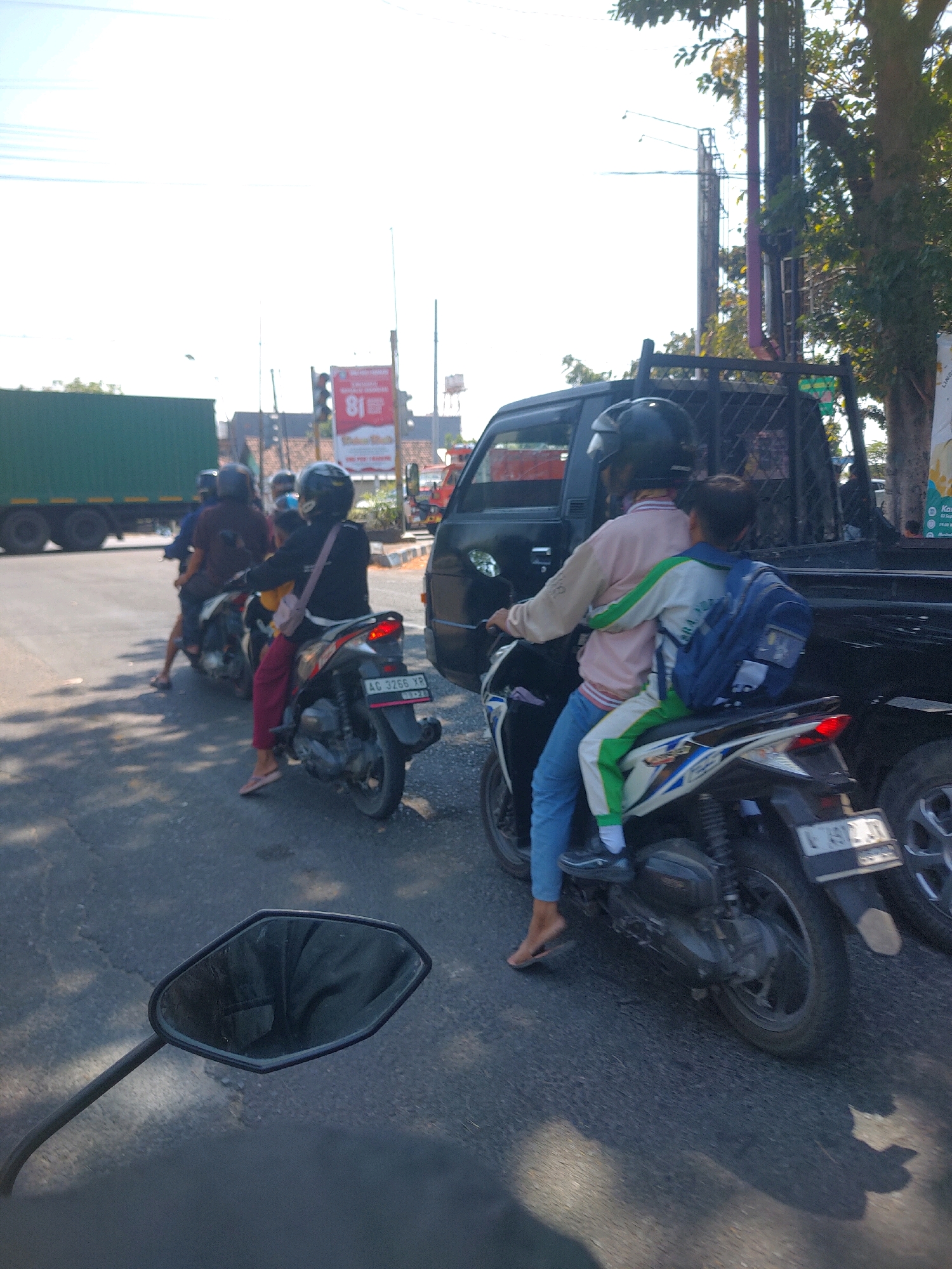 The image depicts a street scene in Begadung, Indonesia, on a sunny day. In the foreground, a motorcycle rear-view mirror is visible. Several motorcycles and a black pick-up truck are on the road, suggesting traffic. On one motorcycle, a driver wearing a helmet and a pink and white jacket is carrying a passenger who has a blue backpack and is wearing a green and white striped outfit. Other motorcycles in the background also carry passengers. A large green shipping container truck is visible to the left, and a traffic light and billboard are present in the background. The road appears to be asphalt.