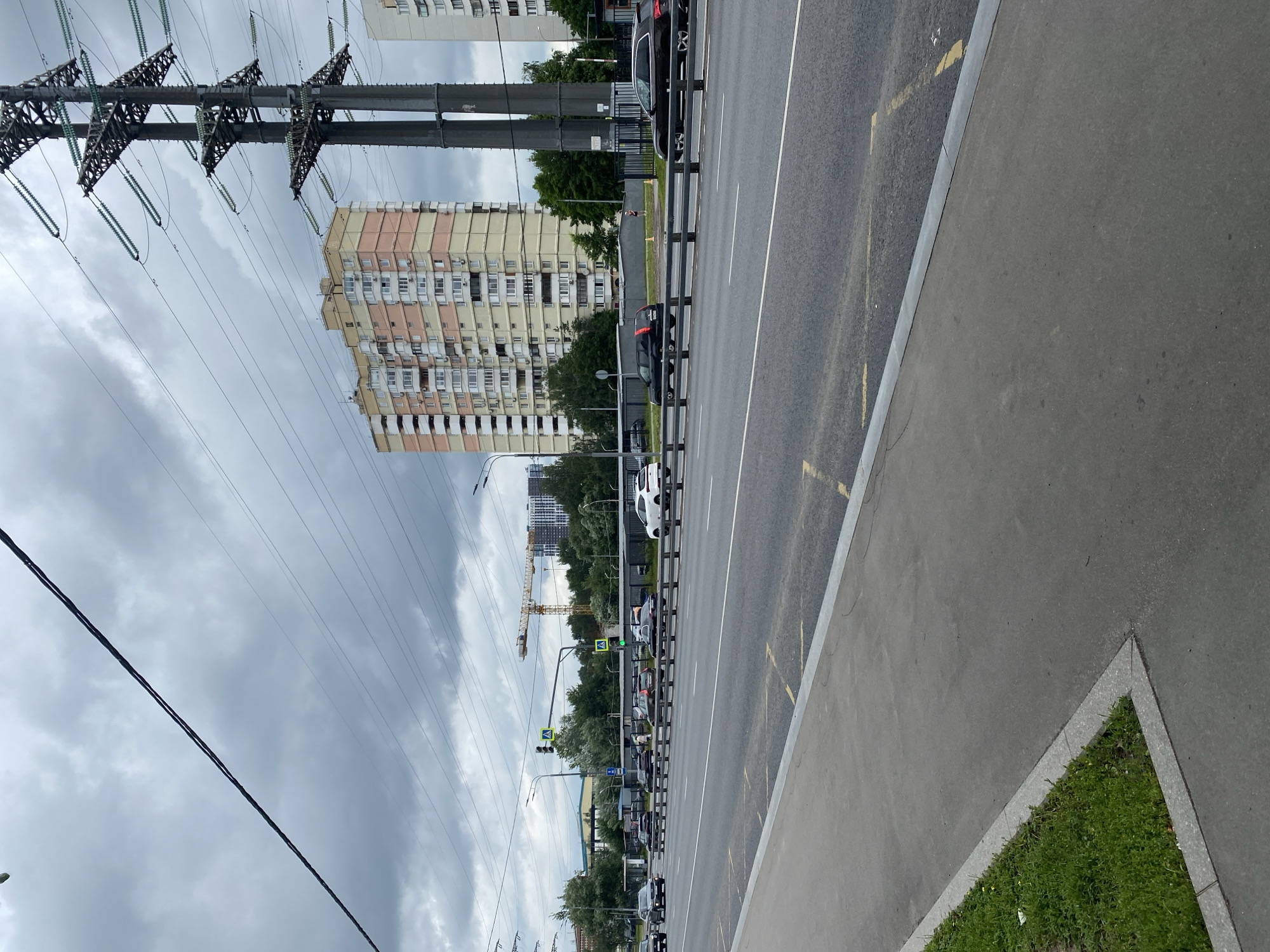 The image depicts a wide, multi-lane road in Khimki, Russia, under a cloudy sky. The foreground shows a grey asphalt sidewalk with a strip of green grass to the left. The road itself has several lanes of traffic with white lane markings and yellow diagonal lines indicating a no-parking zone. A low metal barrier separates the road from the sidewalk area.
Several cars are visible on the road, including a white car in the right lane, a black SUV and a dark sedan in the center lanes, and other vehicles further down the road. Overhead, power lines crisscross the sky, supported by tall, industrial-looking metal towers.
In the background, a multi-story apartment building with a light pink and yellow facade dominates the skyline, featuring numerous windows and balconies. To the right of this building, another, lighter colored apartment building is partially visible. A large construction crane is positioned in the distance, behind trees and other buildings, suggesting ongoing development in the area. Streetlights and traffic signals are also present along the road. The overall impression is of a typical urban environment on an overcast day.