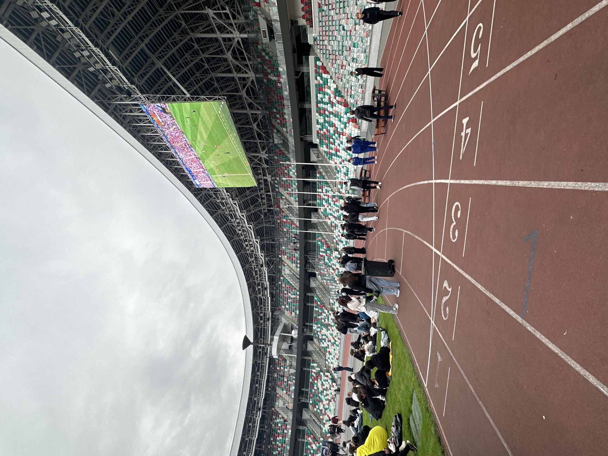 The image shows a partial view of the Dinamo National Olympic Stadium in Minsk, Belarus. The focus is on the running track, with lanes numbered "5" to an obscured lower number. A group of people is gathered on the track and on the infield grass. Some are standing, some are seated on the grass. A large video screen is suspended from the stadium's roof, displaying a scene of a populated stadium, possibly showing a sports event. The stadium seating in the background is a mix of red, green, and white colors. The sky above is overcast. Some people have luggage and are wearing jackets.