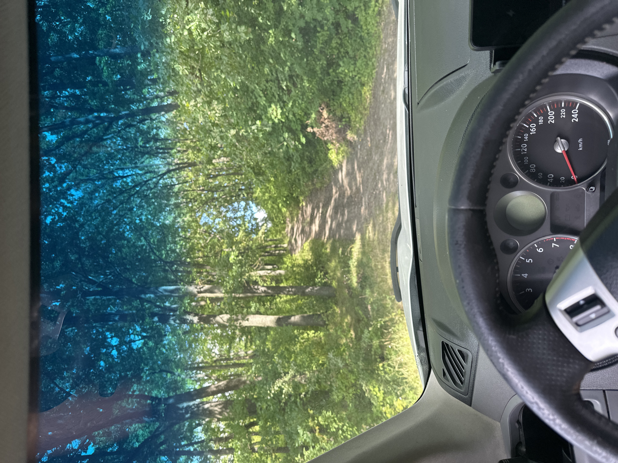 The image shows a view from inside a car, looking out through the windshield. The car is on a dirt road that curves through a dense forest. The trees are tall and green, with sunlight filtering through the leaves. The road is narrow and appears to be unpaved, with loose gravel and dirt. The car's dashboard and steering wheel are visible in the foreground, indicating the perspective of the driver. The windshield has a blue tint, common for UV protection. The environment suggests a rural or natural setting, possibly a park or a less-traveled road outside of a populated area. There are no people visible in the image. The scene appears to be captured during daylight hours on a clear day. The location is stated to be Shatava, Ukraine, which aligns with the rural, forested environment depicted.