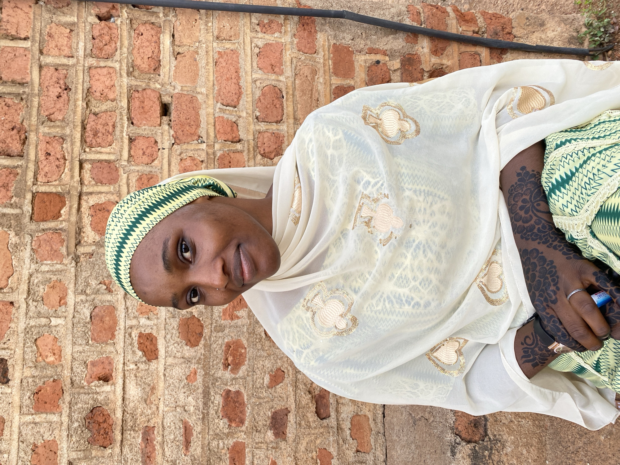 A woman stands near a brick wall. She is wearing a white headscarf decorated with gold and green accents and a yellow and green cap. The background features a brick wall, typical of buildings in Vom, Nigeria, and a black metal pipe is visible at the top. The woman is wearing a ring and has henna tattoos on her hand.