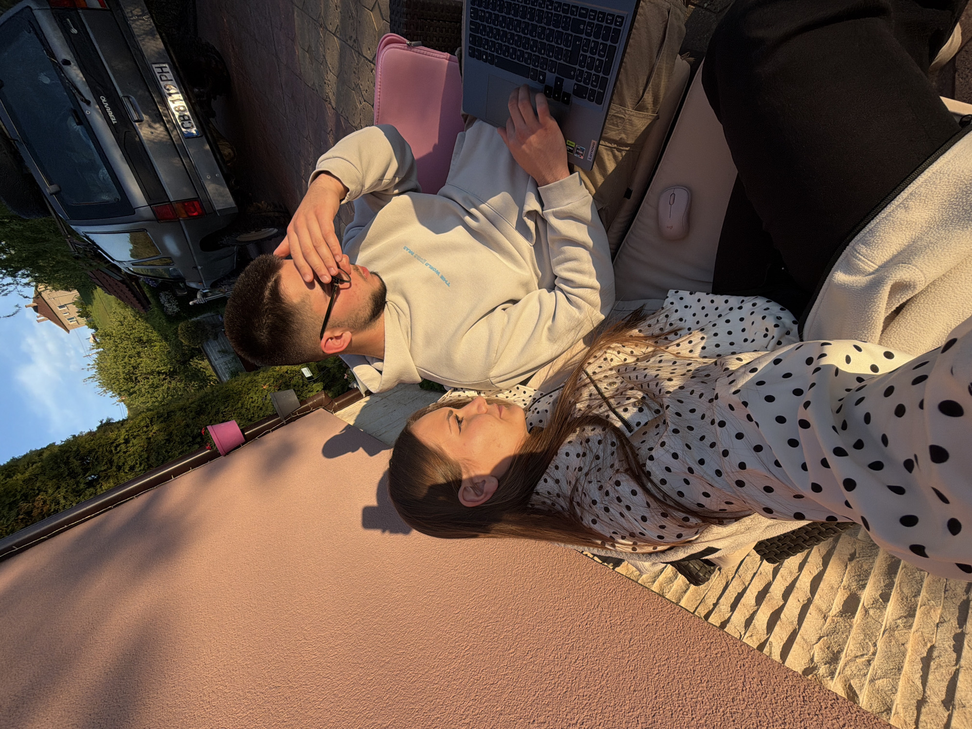 A man and a woman are lying on a blanket in Buhovo, Bulgaria. The man is lying on his back with a laptop on his lap. He is wearing a light-colored hooded sweatshirt and shorts. The woman is lying next to him, wearing a polka-dotted dress. A gray car is parked in the background.