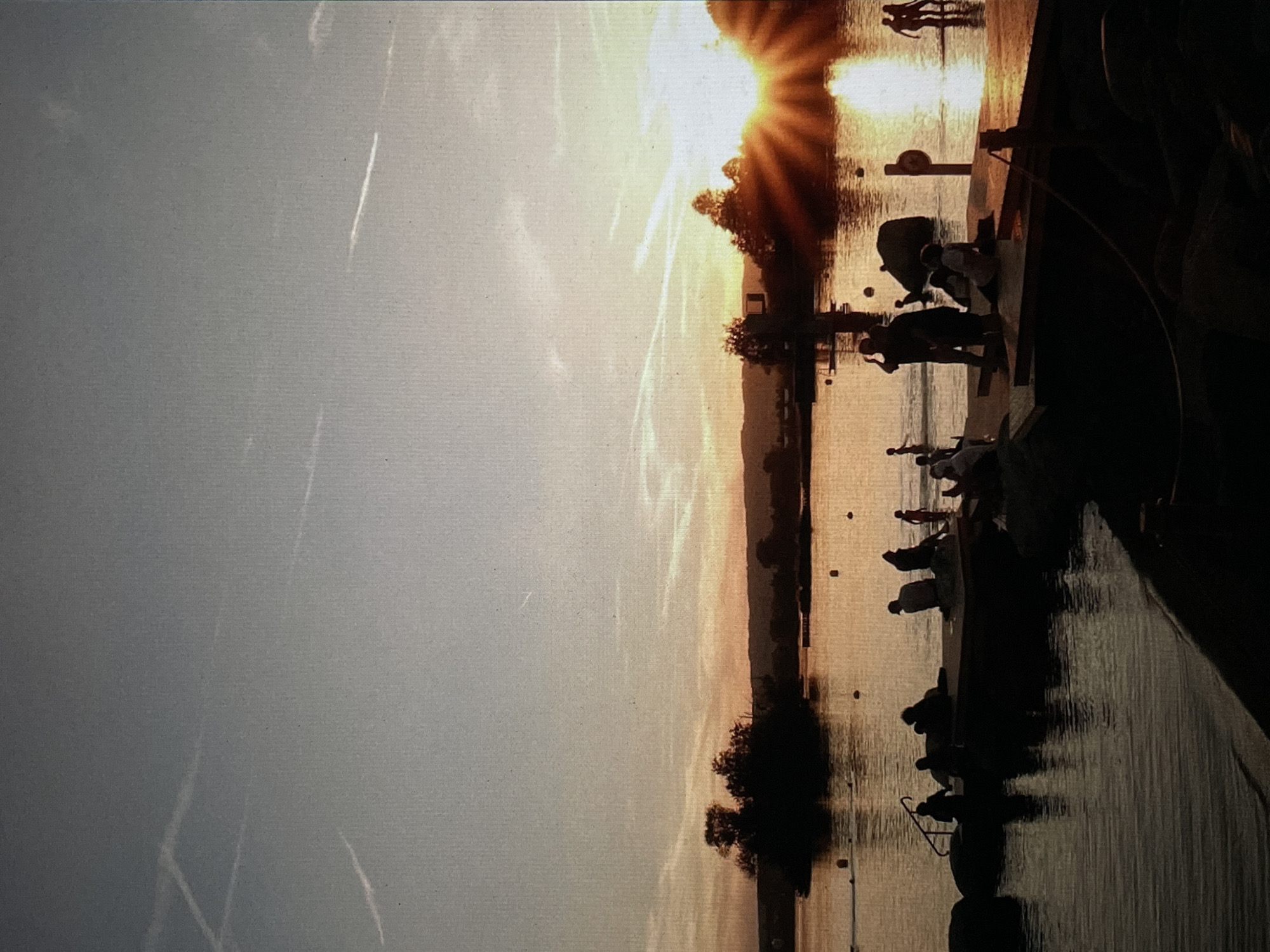 The image depicts a sunset scene over water, likely a river or bay, with the sun's rays visible through trees. The scene is silhouetted against the bright sky, revealing figures of people standing along a waterfront. The silhouette suggests trees are present in the background, and some structures can be discerned on the water's edge. Vapor trails are visible in the sky. Based on the information available, I am unable to ascertain this is in Saint Petersburg, Russia.