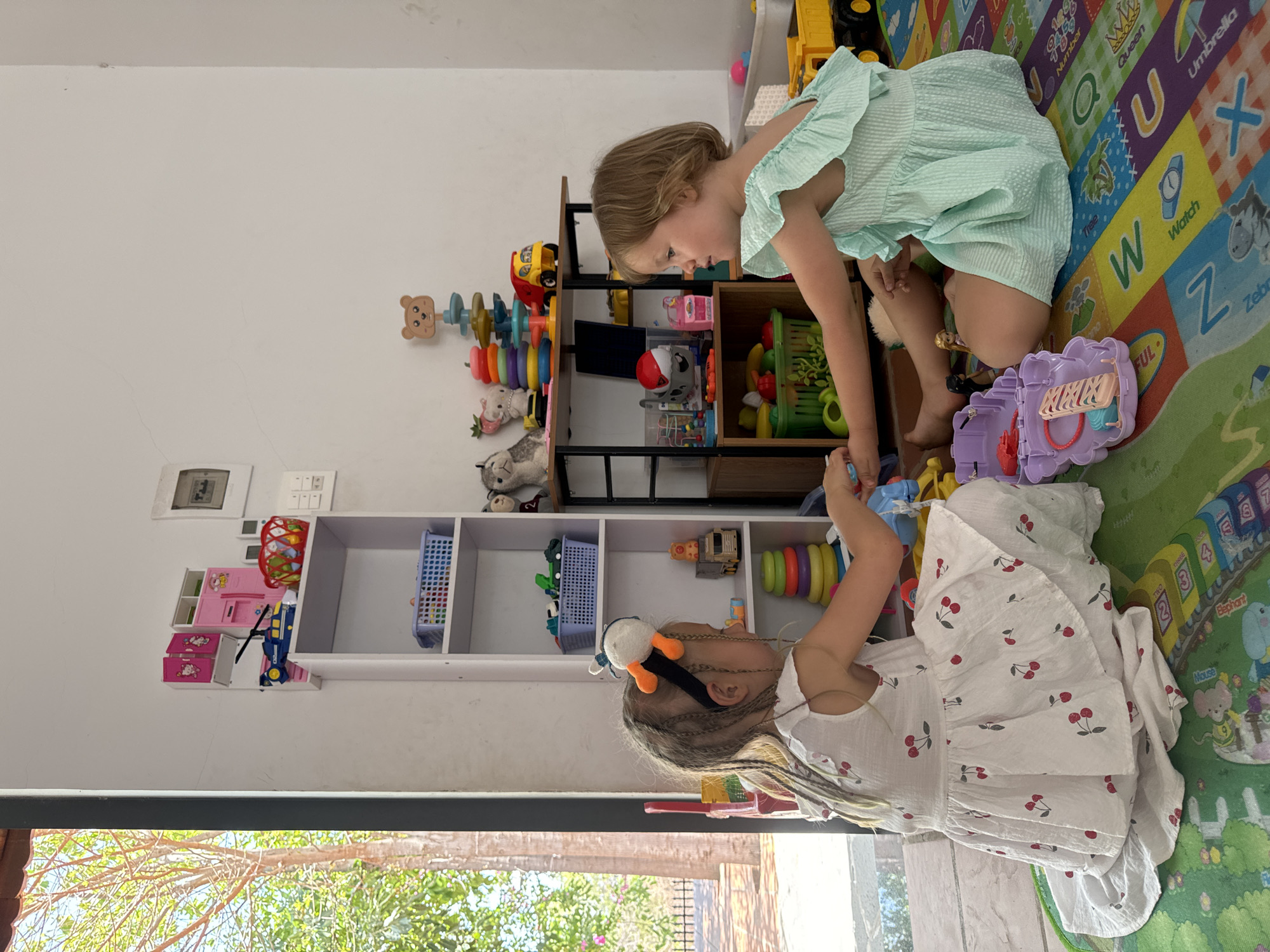 Two young girls are playing in a room full of toys. The girl on the right wears a light blue dress and leans over a small dark wooden shelving unit with colorful toys on it. The girl on the left wears a white dress with red cherry print and faces the girl on the right. The girls are on a colorful ABC rug. The floor is light-colored tile. To the left, on a white wall, is a light switch and a small shelf with pink and blue toys. There is a window near the bottom of the image showing a lush green outdoor area. This scene could be located in Mũi Né, Vietnam.