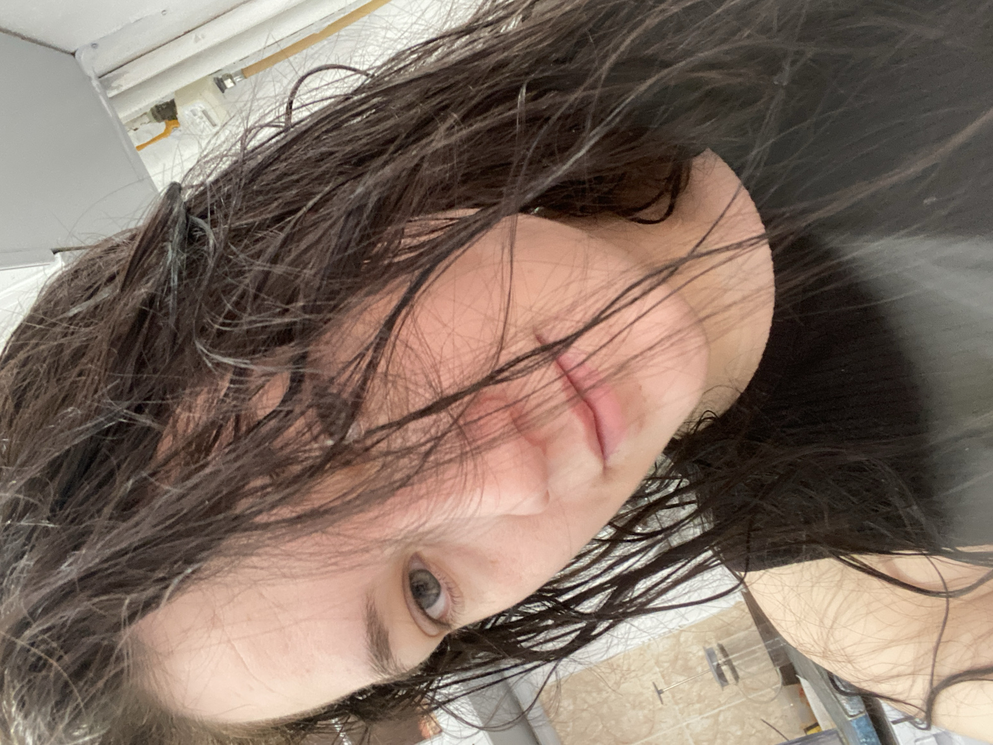 A selfie-style photograph depicts a woman with wet, dark hair. Her face is partially obscured by strands of hair, but a light-colored complexion, a single visible eye with light pigmentation, and a portion of her lips are discernible. The background includes ceiling and wall elements suggestive of an interior space, along with some indistinct plumbing fixtures. The overall image orientation is tilted. There is no explicit information to locate the subject in Chaikovsky, Russia.