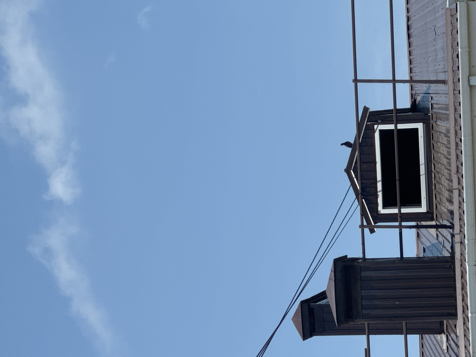 A low-angle, close-up outdoor shot captures a section of a rooftop against a clear blue sky with sparse white clouds, likely taken in Mikhaylovsk, Russia. Dominating the foreground are two dark brown, rectangular chimney structures with tapered caps, standing tall on a dark brown, corrugated metal roof. A metal railing runs horizontally across the mid-ground, forming a barrier. To the right of the chimneys, a dormer window with a white frame is visible, its opening completely black, suggesting it is either unglazed or heavily shaded. Perched on the peak of the dormer is a lone black bird, silhouetted against the sky. Power lines are visible stretching diagonally across the upper left portion of the image. The overall impression is one of urban infrastructure against a bright, open sky.