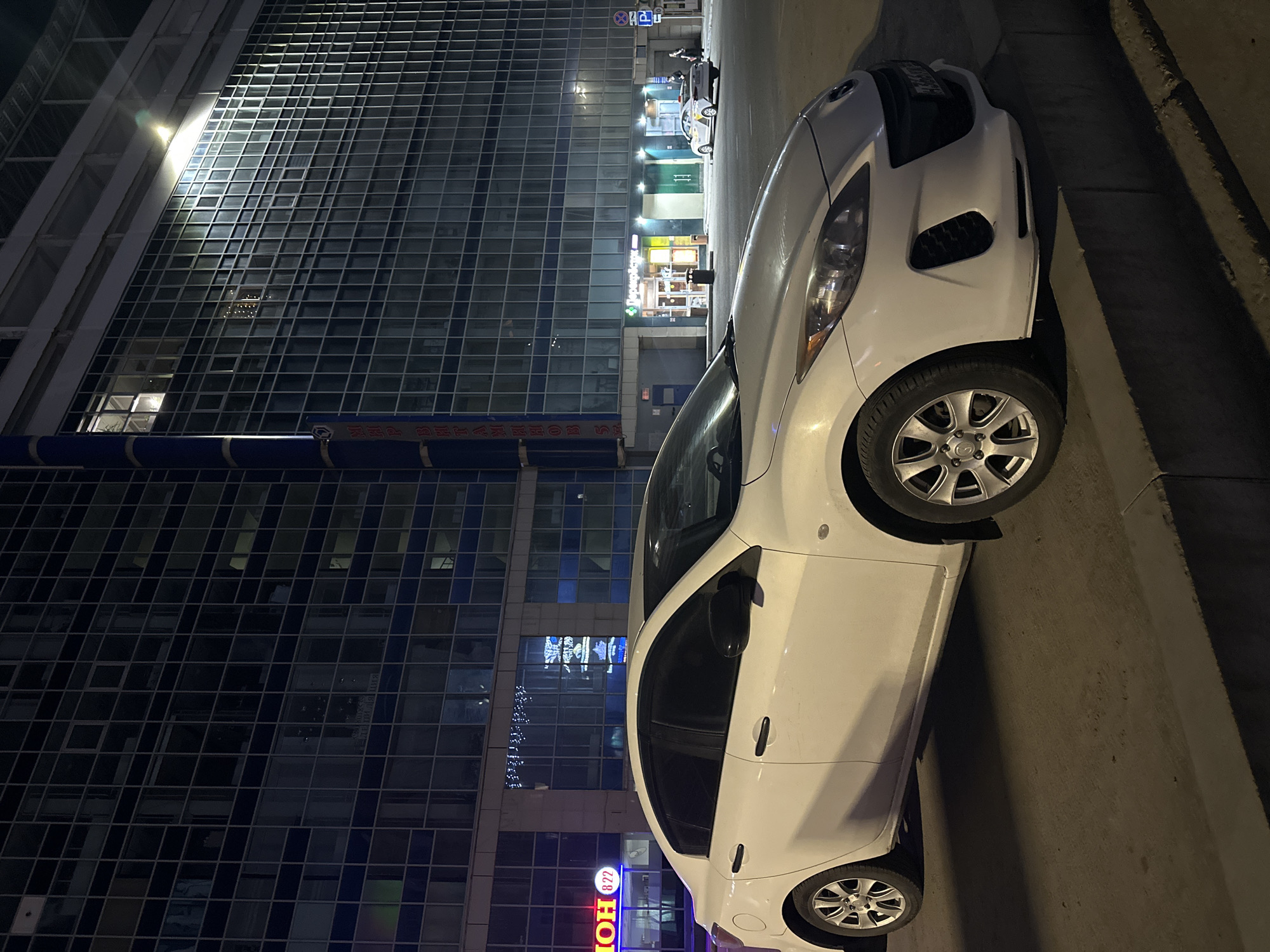 The image depicts a white compact car parked on a city street. The backdrop reveals a modern glass-fronted building, illuminated by interior lights and street lamps, reflecting the urban environment.
The sign "MOH 822" is visible at the base of the building. Based on the Cyrillic lettering and overall urban setting, the location is plausibly Perm, Russia.
There is no explicit indication of people in the image. The focus is on the parked car and the cityscape. The setting is an urban street at night, as evidenced by the dark ambient light and artificial lighting from the building and street lights.