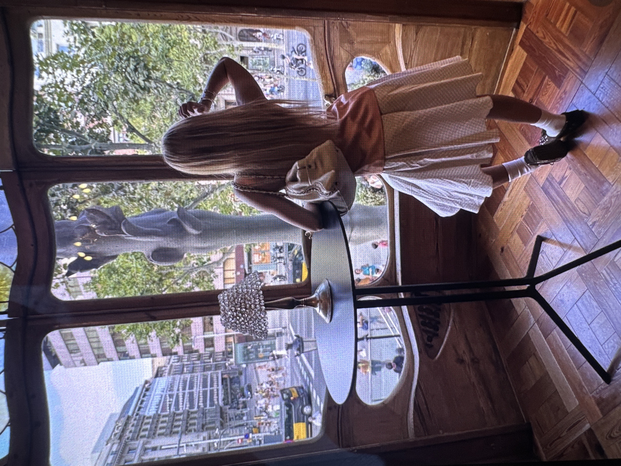 A woman with long blond hair wearing a white dress and carrying a shoulder bag is standing by a table with a vase on it. She is looking out the window at a city street with buildings and trees. In the background, there is a statue. The location is Unknown, Unknown. The floor is wooden.