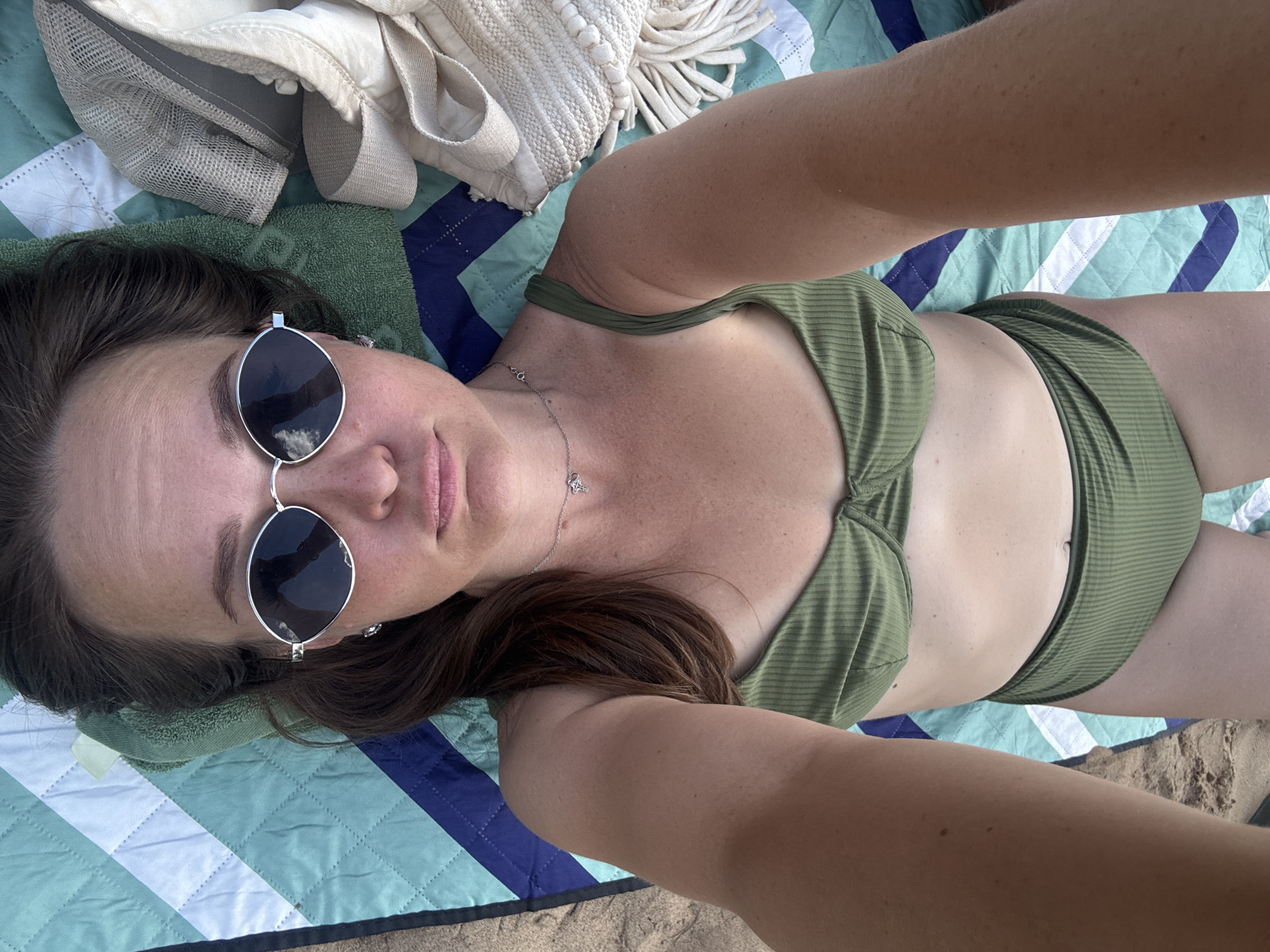 The image shows a woman lying on a beach blanket. She is wearing a green bikini and sunglasses. There is a towel and a bag next to her. The woman is looking at the camera. The background is a beach with sand and water. The specific location in the city of Novyi, Russia, is not visible in the image.
