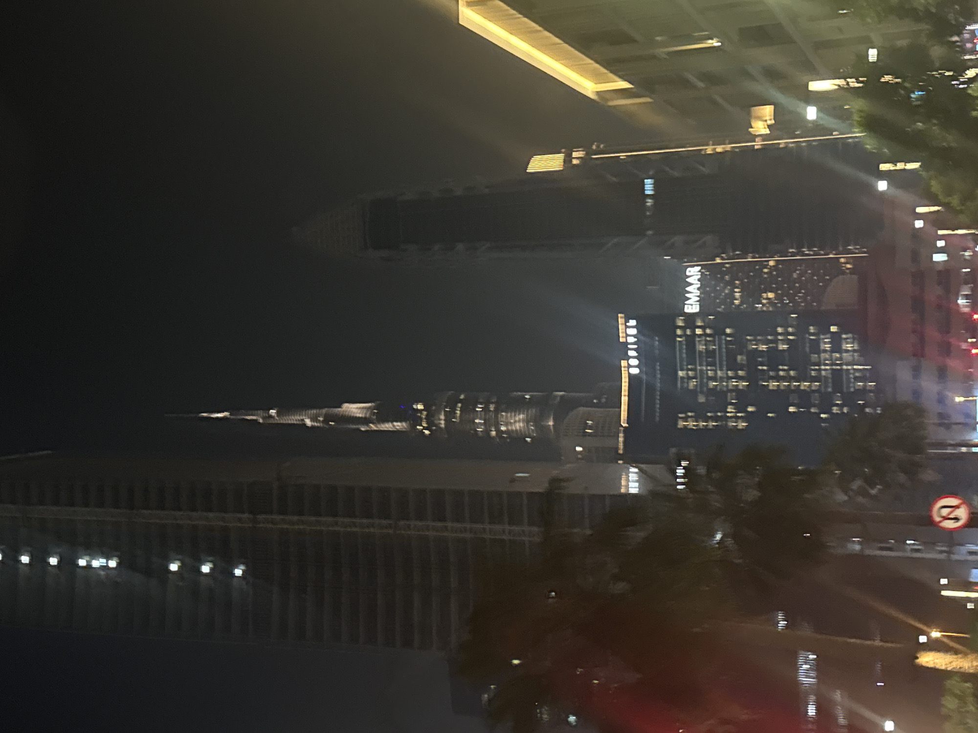 The image shows a nighttime view of the Burj Khalifa and surrounding skyscrapers in Dubai, United Arab Emirates. The Burj Khalifa is illuminated and stands prominently in the center-right of the frame. To its left, another tall skyscraper is visible, under construction or with some lights on. In the foreground, palm trees obscure parts of the lower sections of the buildings, and a traffic sign indicating a "no U-turn" is visible on the right. Other buildings, including one with "EMAAR" signage, and streetlights are also present, contributing to the urban night scene. The sky is dark, indicating it is nighttime, and there appears to be some haze or fog contributing to the diffused lighting.