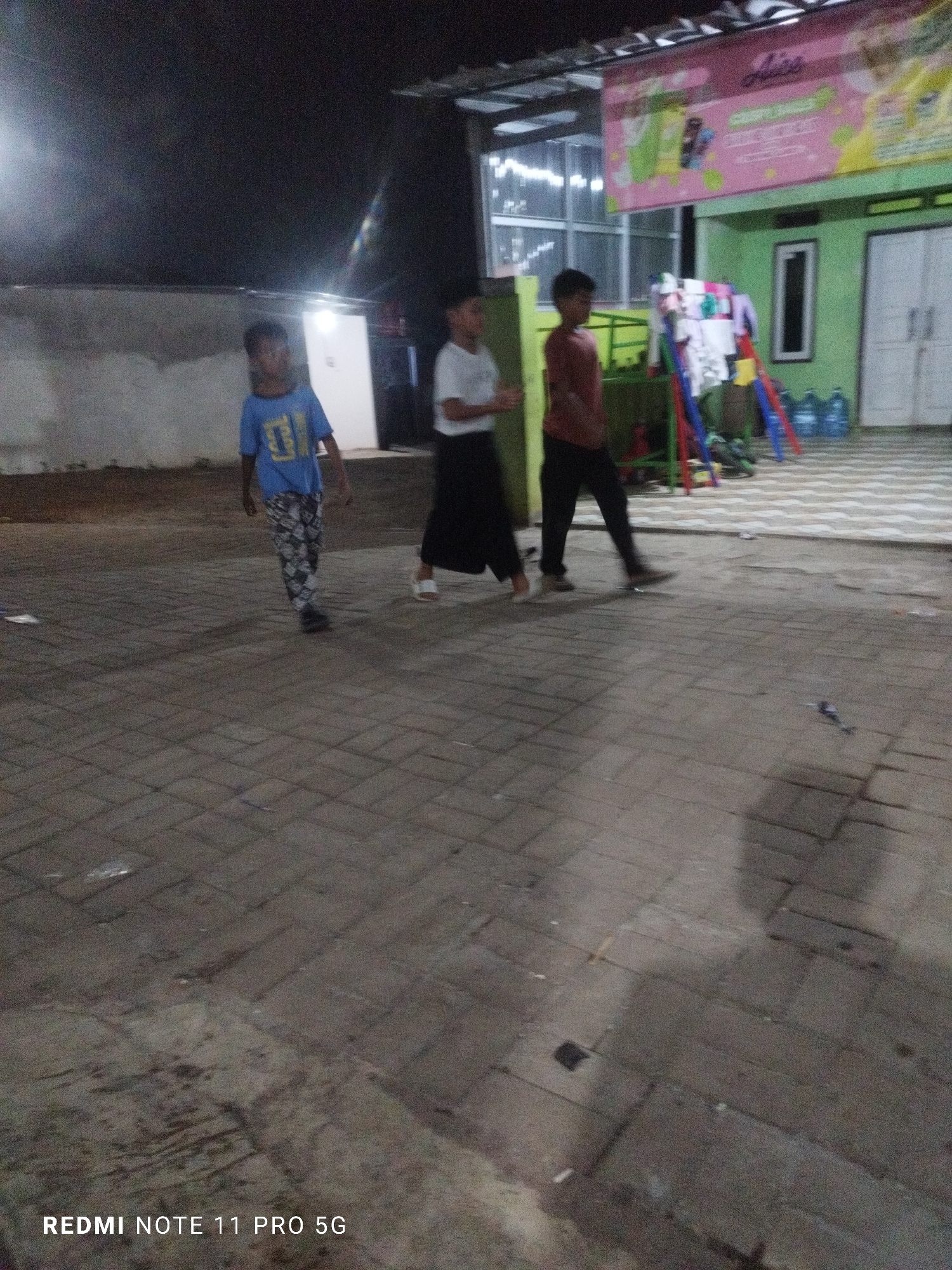 The image depicts an outdoor scene at night, likely in Kaduhejo, Indonesia. Three boys are walking on a paved area. The boy on the left wears a blue t-shirt and patterned pants. The boy in the middle wears a white t-shirt and dark pants. The boy on the right wears a reddish-brown t-shirt and dark pants. Behind them, a building with green walls and a pink banner is visible. There are also what appear to be large blue water bottles to the right of the building. The ground is paved with interlocking stones.