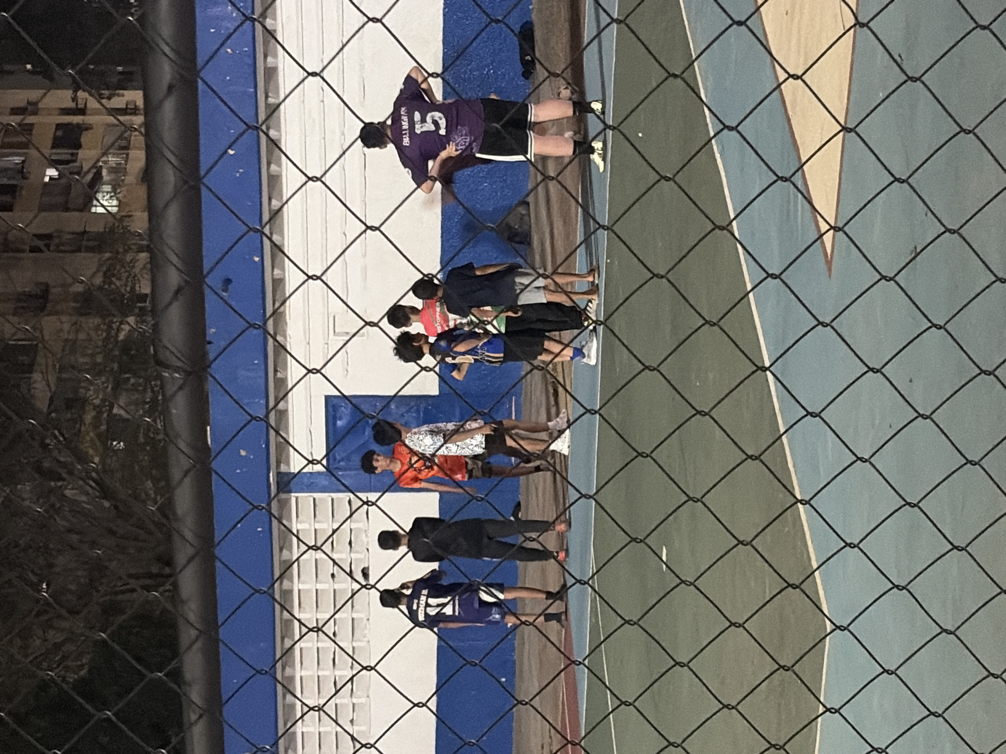 The image shows a group of people standing on an outdoor court behind a chain-link fence. The court has a blue and green surface with white markings. A wall with blue and white horizontal stripes is behind the group. One person is wearing a purple jersey with the number "5" and the name "BELLWARD" on the back. Other individuals are wearing various athletic attire. The scene appears to be at night, with some artificial lighting illuminating the area. In the background, there's a glimpse of buildings, possibly residential. Given the context clues, the location is likely in Ciudad Guayana, Venezuela.