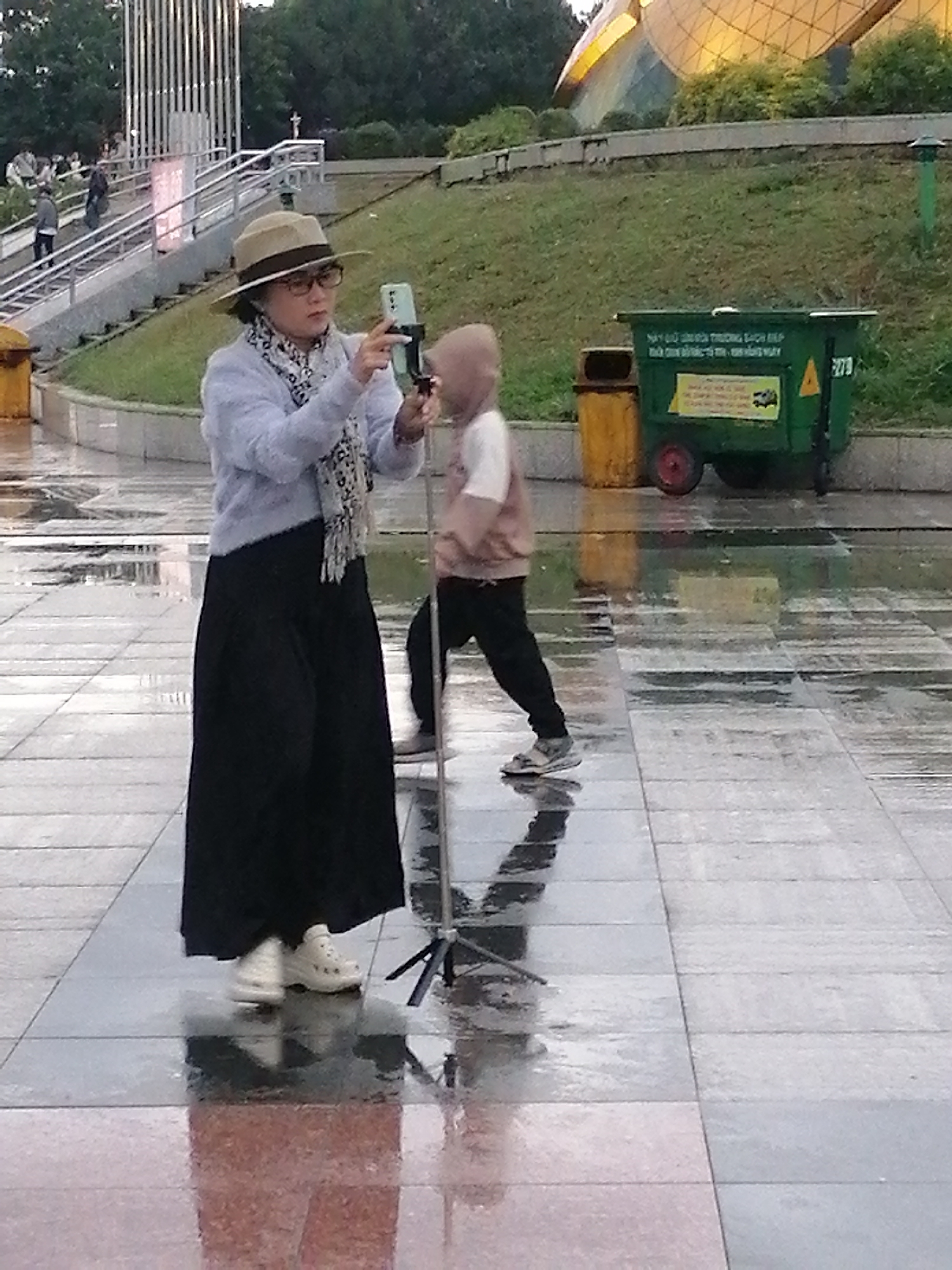 The image shows an outdoor scene in Da Lat, Vietnam, characterized by wet, reflective paving stones, suggesting it might be raining or recently rained. In the foreground, a woman wearing a wide-brimmed hat, glasses, a light blue sweater, and a long black skirt is holding a smartphone on a selfie stick, appearing to take a picture or video. She has a patterned scarf around her neck. To her right and slightly behind her, a child wearing a light brown hooded jacket and dark pants is walking away from the camera. In the background, there are grassy slopes and a distinctive building with a golden, curved roof, possibly a landmark. Further back, a green trash bin with wheels and a yellow trash can are visible. A set of stairs with a metal railing leads up a slope. Other people can be seen in the distance on the stairs and walking. The overall atmosphere is that of a public space in a city, with urban elements and natural landscaping.