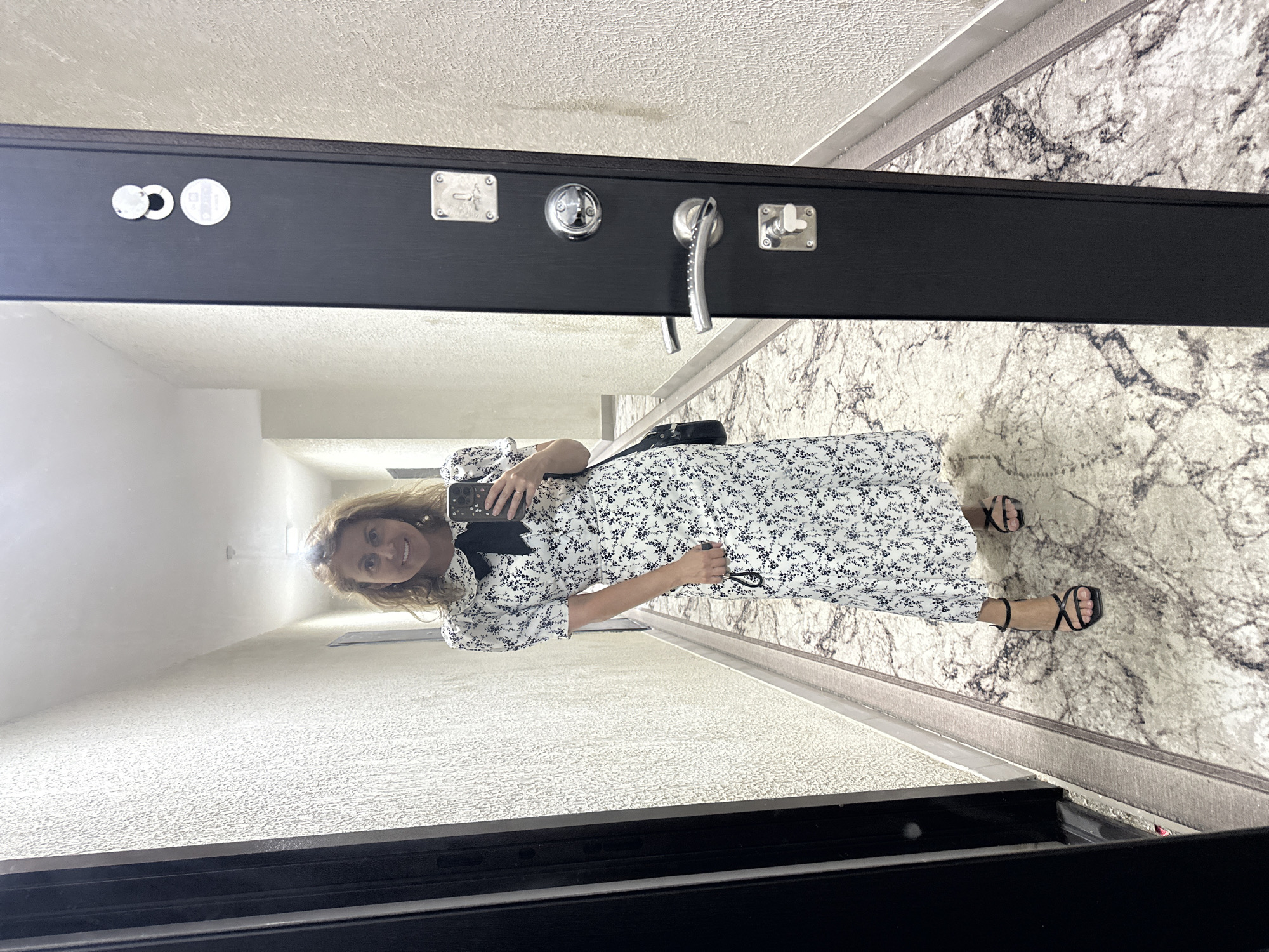 A woman stands in a hallway and takes a selfie in a full-length mirror. She is wearing a white dress with a black floral print, a black bow tie, and black high-heeled sandals. She is holding a black object in her right hand and a smartphone in her left hand. The hallway has textured walls and a carpeted floor. A black door with silver hardware is visible on the right side of the image. The location is identified as Verkhniy Yurt, Russia.